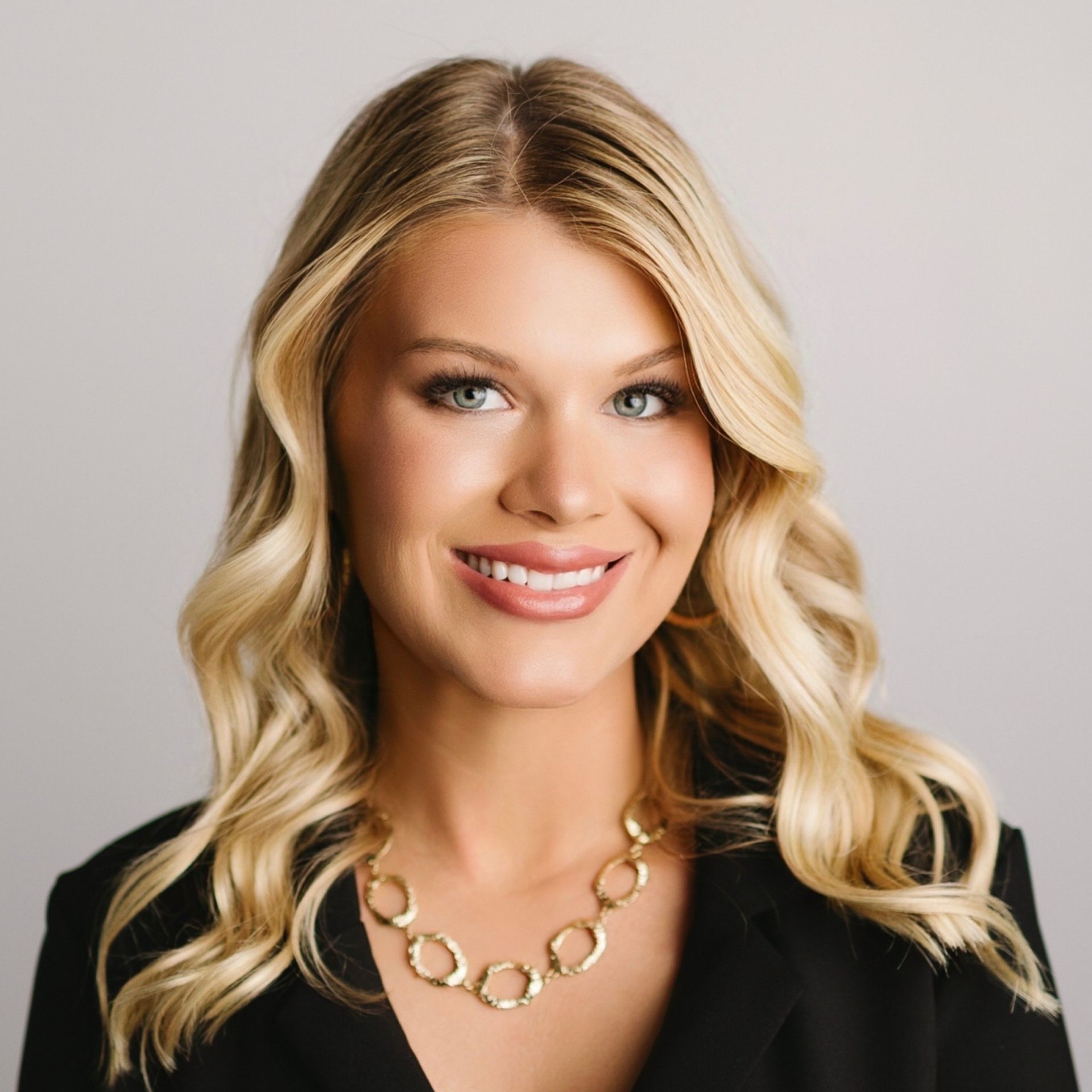 Woman with blonde hair wearing a black blazer and gold necklace, smiling, against a light gray background.