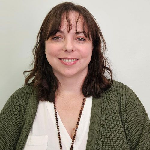 Woman with shoulder-length brown hair, smiling, wearing a white shirt and olive green cardigan, with a beaded necklace.