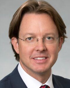 Man with light brown hair, glasses, and a blue suit smiling at the camera.