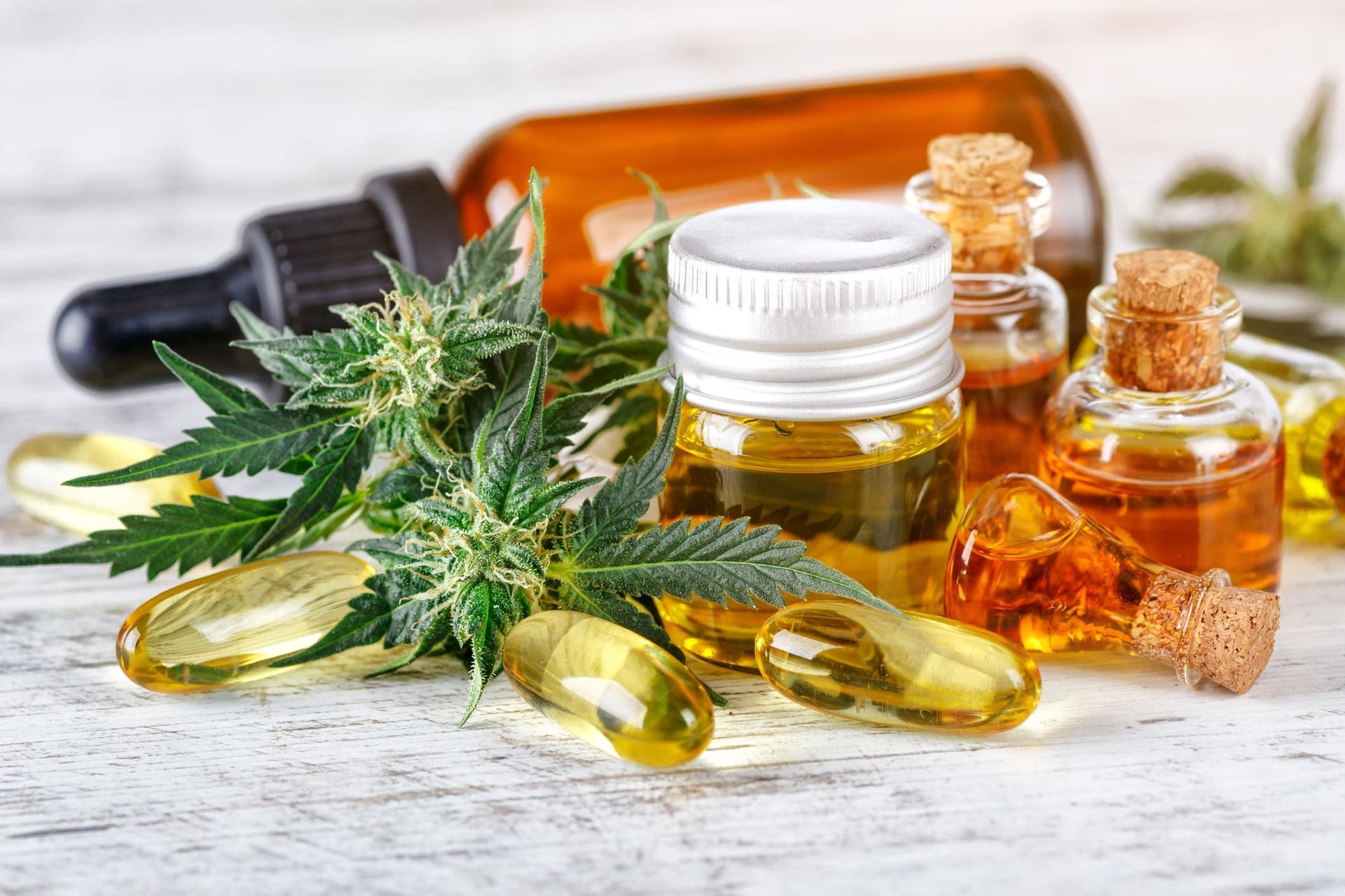 Cannabis plant with buds, oil bottles, and capsules on a white wooden surface.