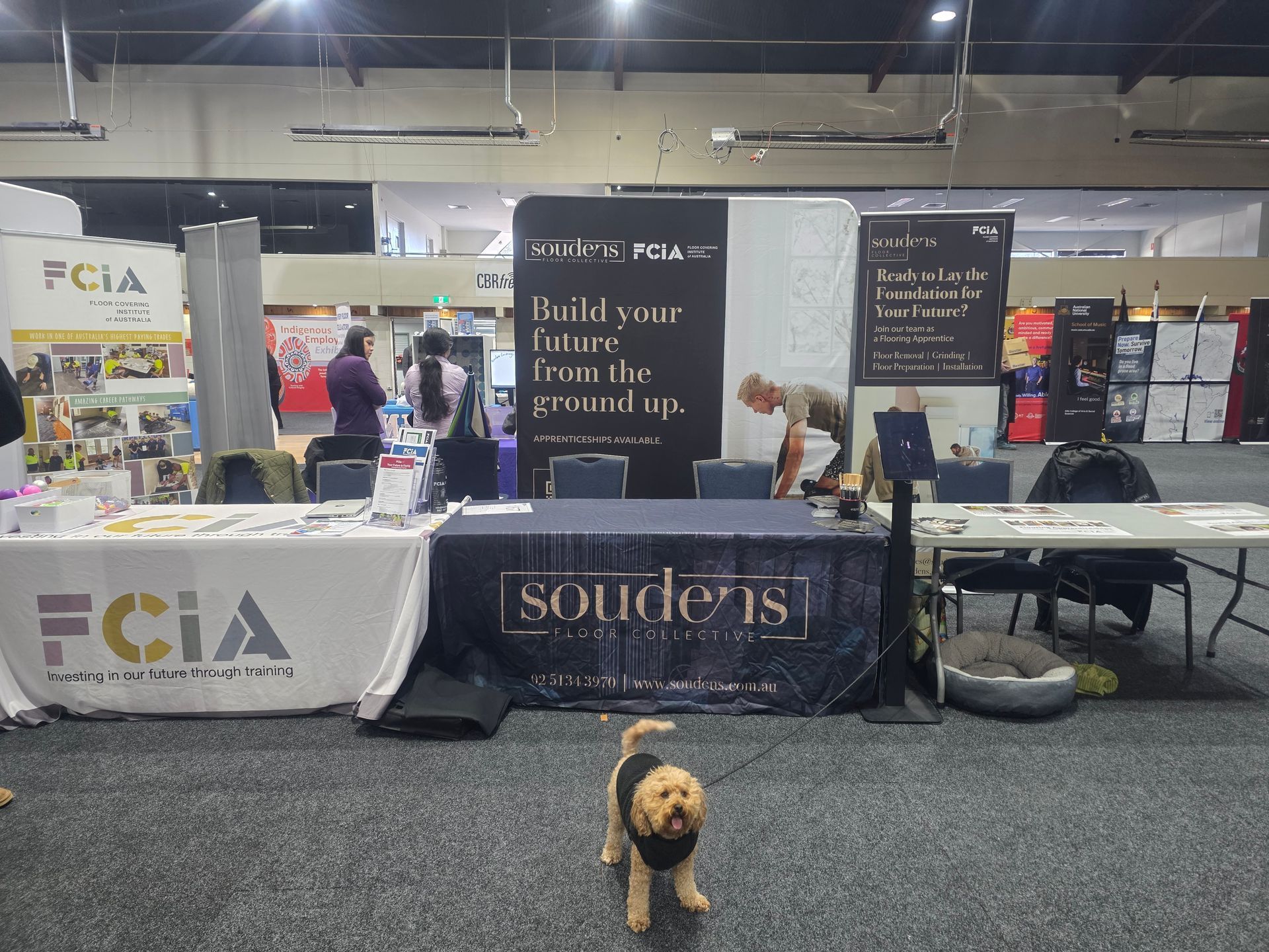 Trade show booth for “souders” with banners, tables, and a small dog standing in front of it.