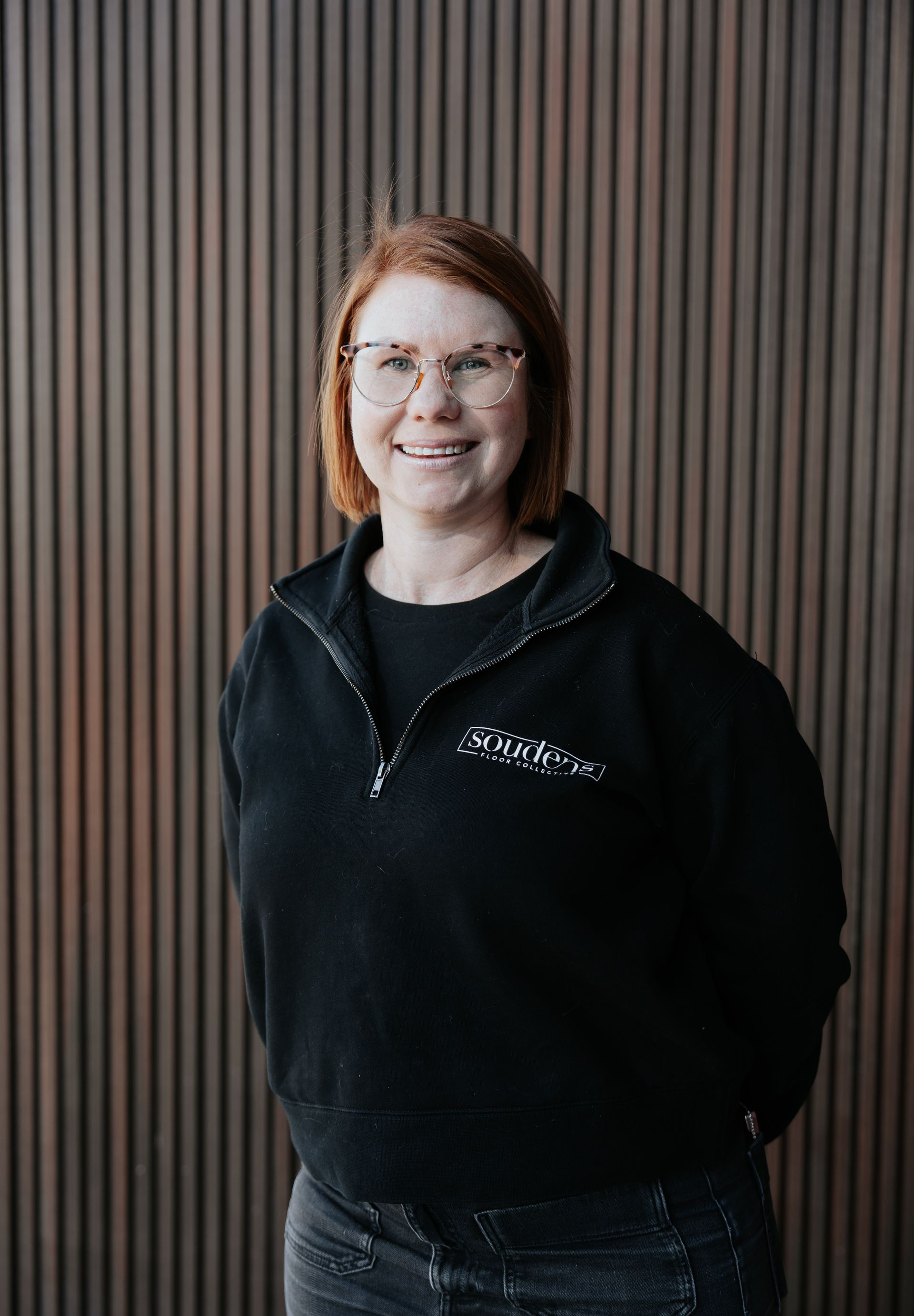 Woman with red hair and glasses smiles, wearing black sweater, standing in front of brown wall.