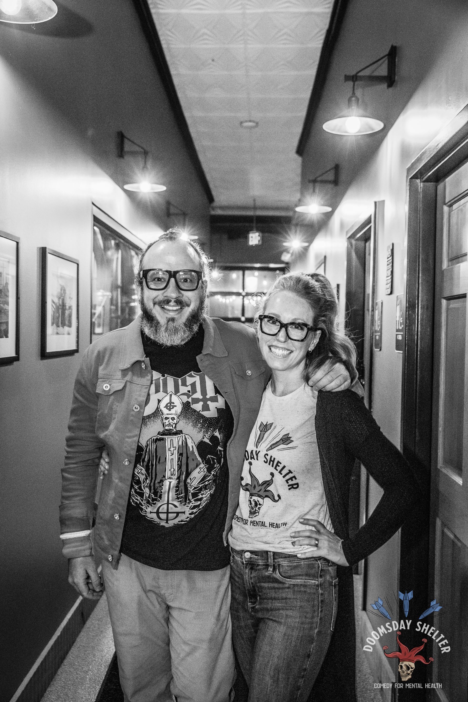 Two people, wearing glasses, stand close in a narrow hallway. Man with tattoos, woman with hand on hip.