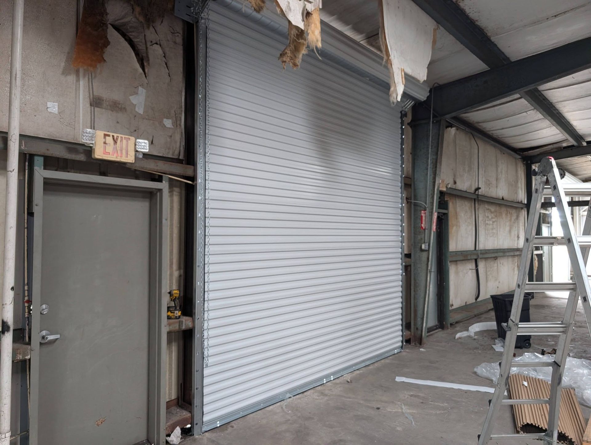 Interior view of a building under construction. A large gray roll-up door is closed next to a closed gray door.