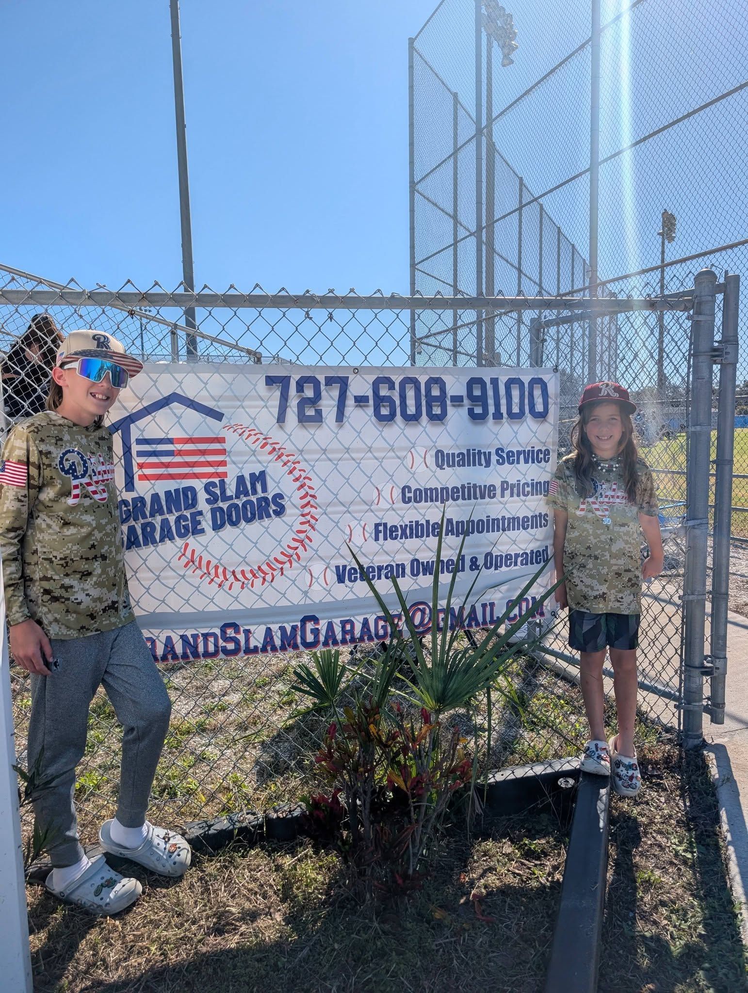 Two children stand near a chain link fence with a Grand Slam Garage Doors sign. They wear matching camouflage shirts. Two children stand near a chain link fence with a Grand Slam Garage Doors sign. They wear matching camouflage shirts.