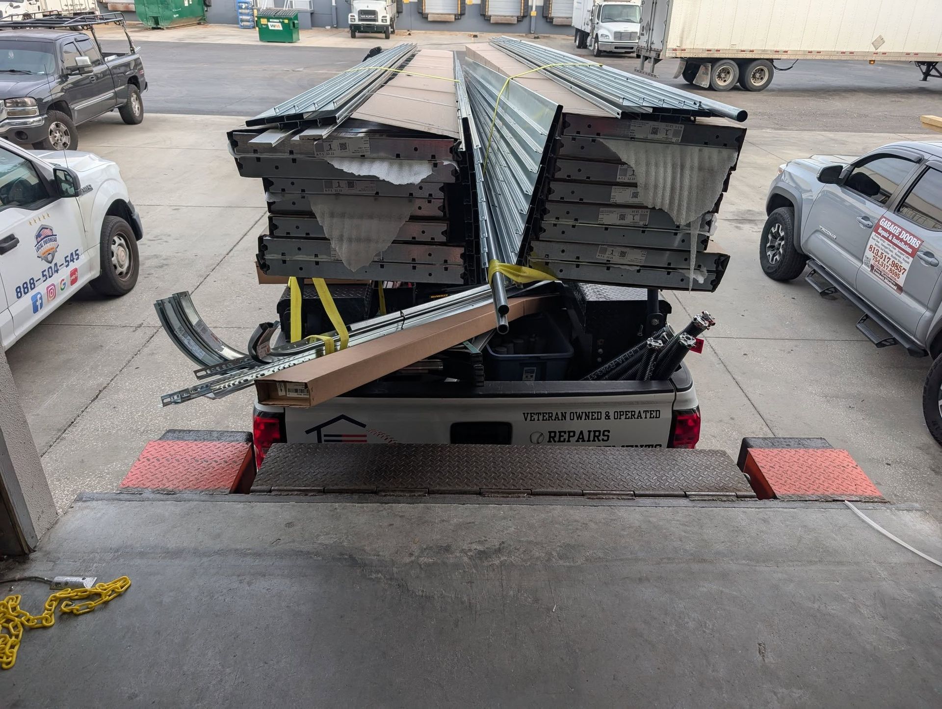 Truck bed loaded with metal components at a loading dock. Two trucks visible.