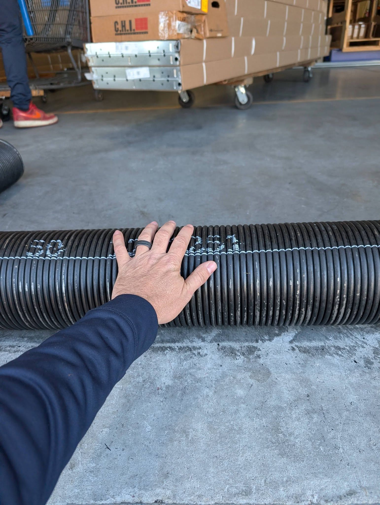 Hand on a large, black, corrugated hose on a concrete floor in a warehouse setting. Hand on a large, black, corrugated hose on a concrete floor in a warehouse setting.