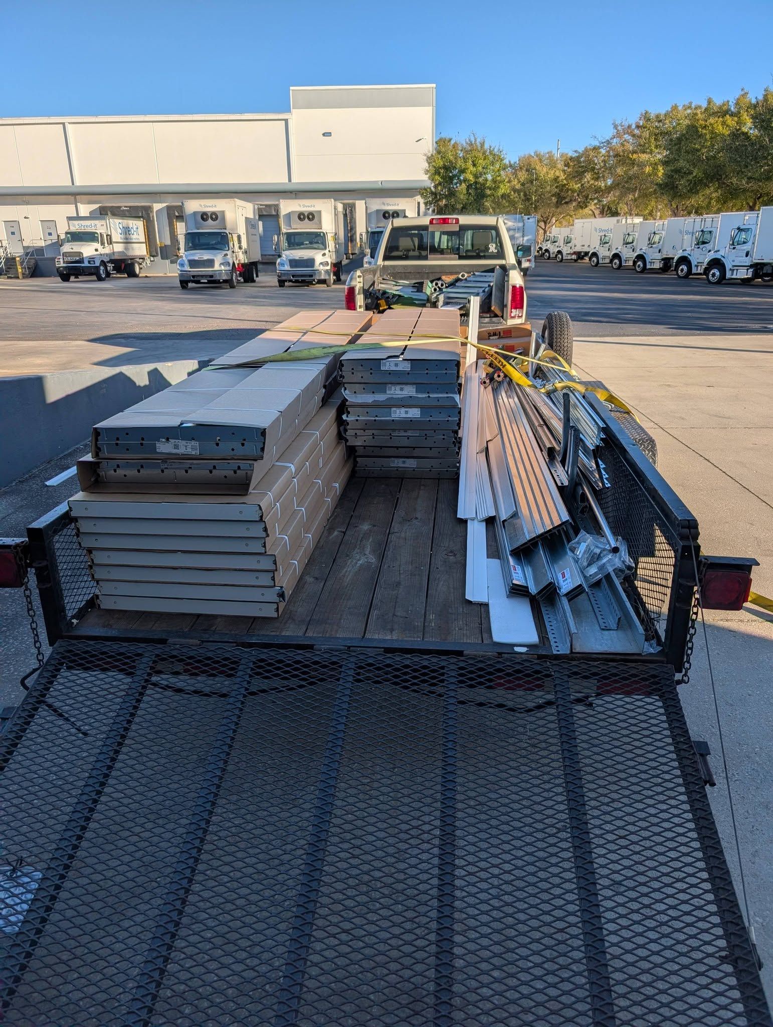 Trailer loaded with construction materials, parked in a lot. White truck in the background, other vehicles visible. Trailer loaded with construction materials, parked in a lot. White truck in the background, other vehicles visible.
