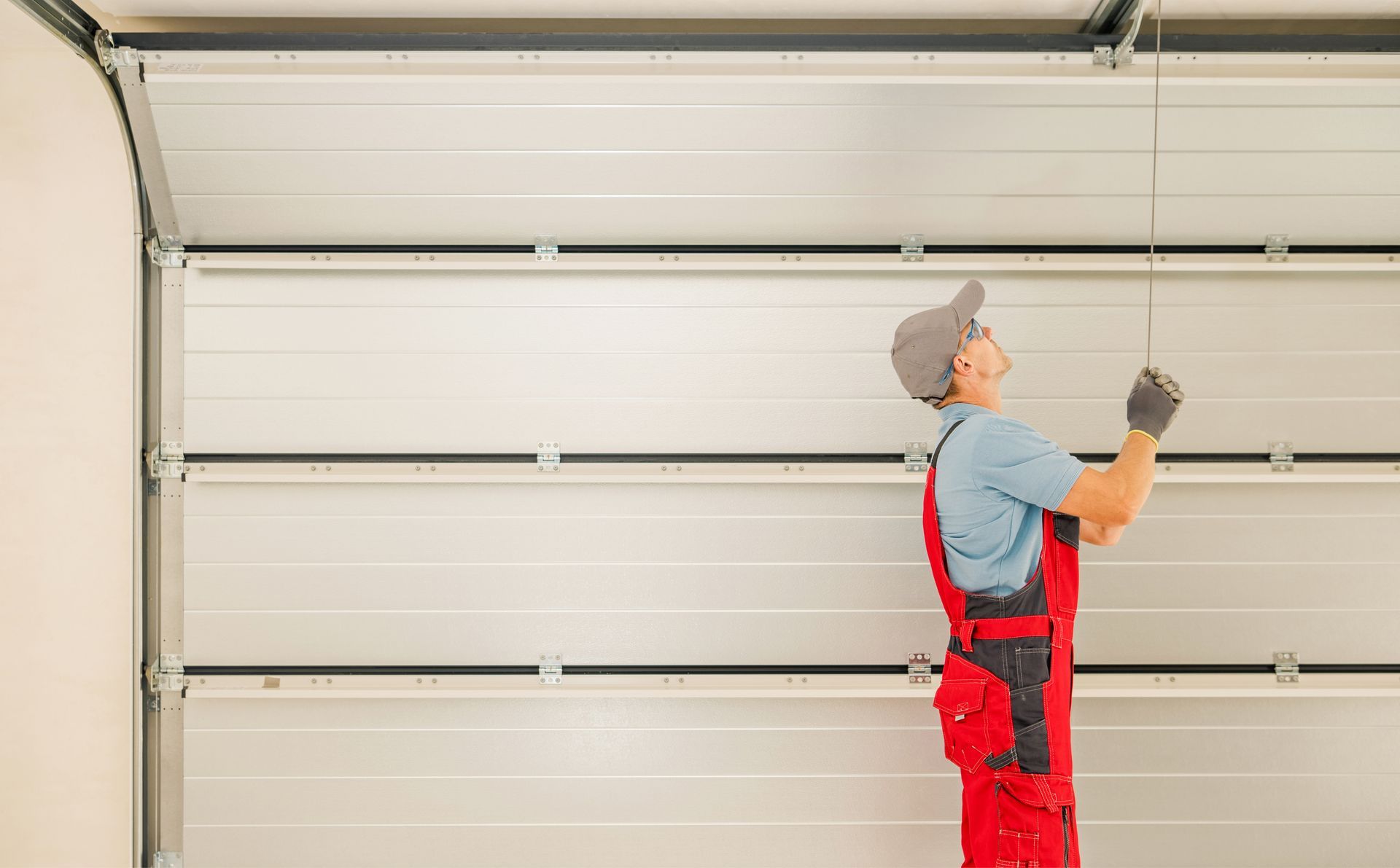 Man in red overalls repairing a garage door. He's looking up, pulling a cable. Man in red overalls repairing a garage door. He's looking up, pulling a cable.
