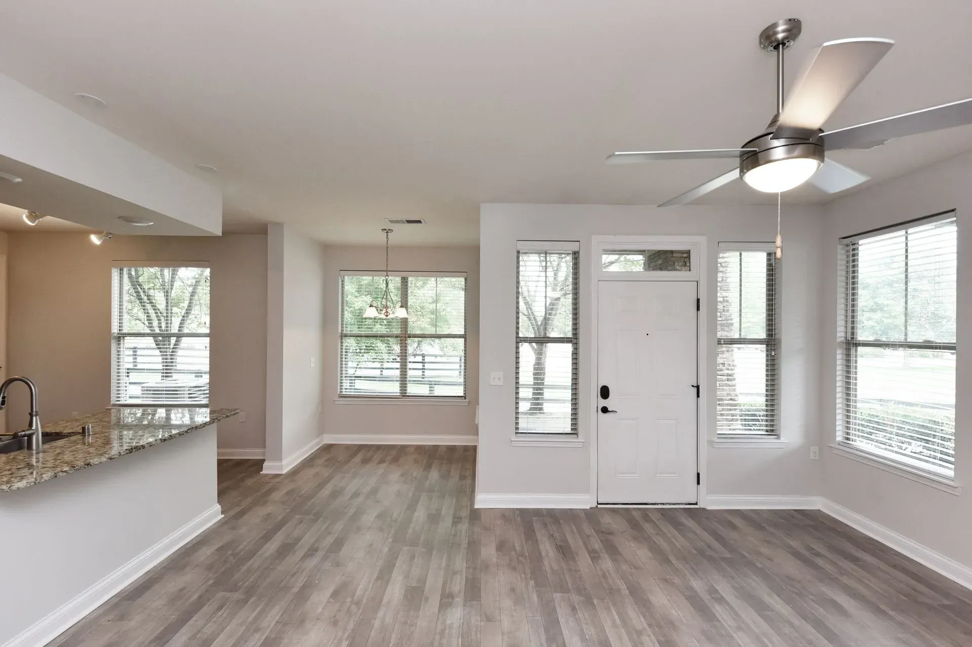 Interior view of an empty apartment living room, dining room, and kitchen area with wood-look flooring and a ceiling fan.