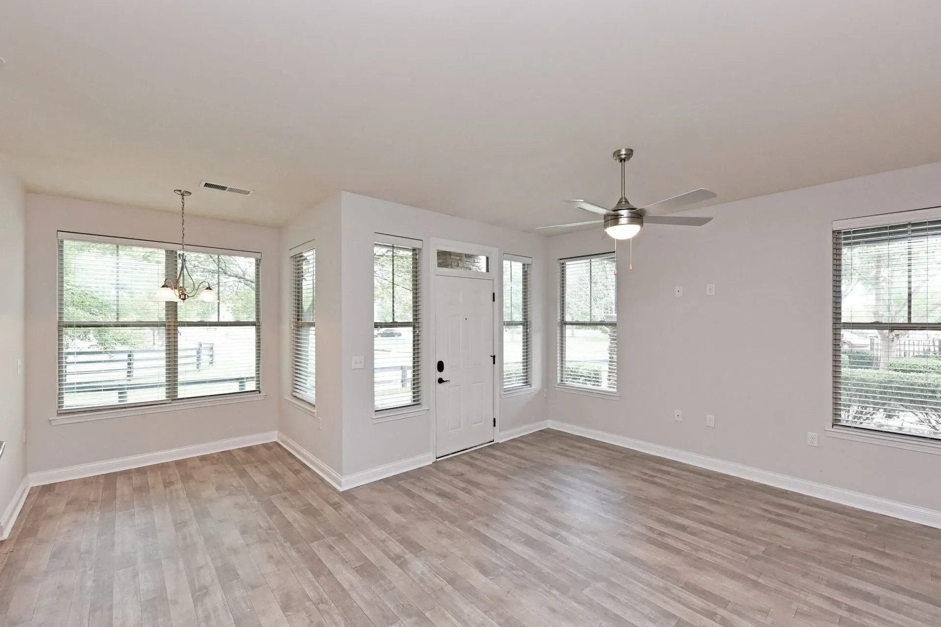 Interior view of an empty apartment living room with wood-look flooring, a white front door, large windows, and a ceiling fan.