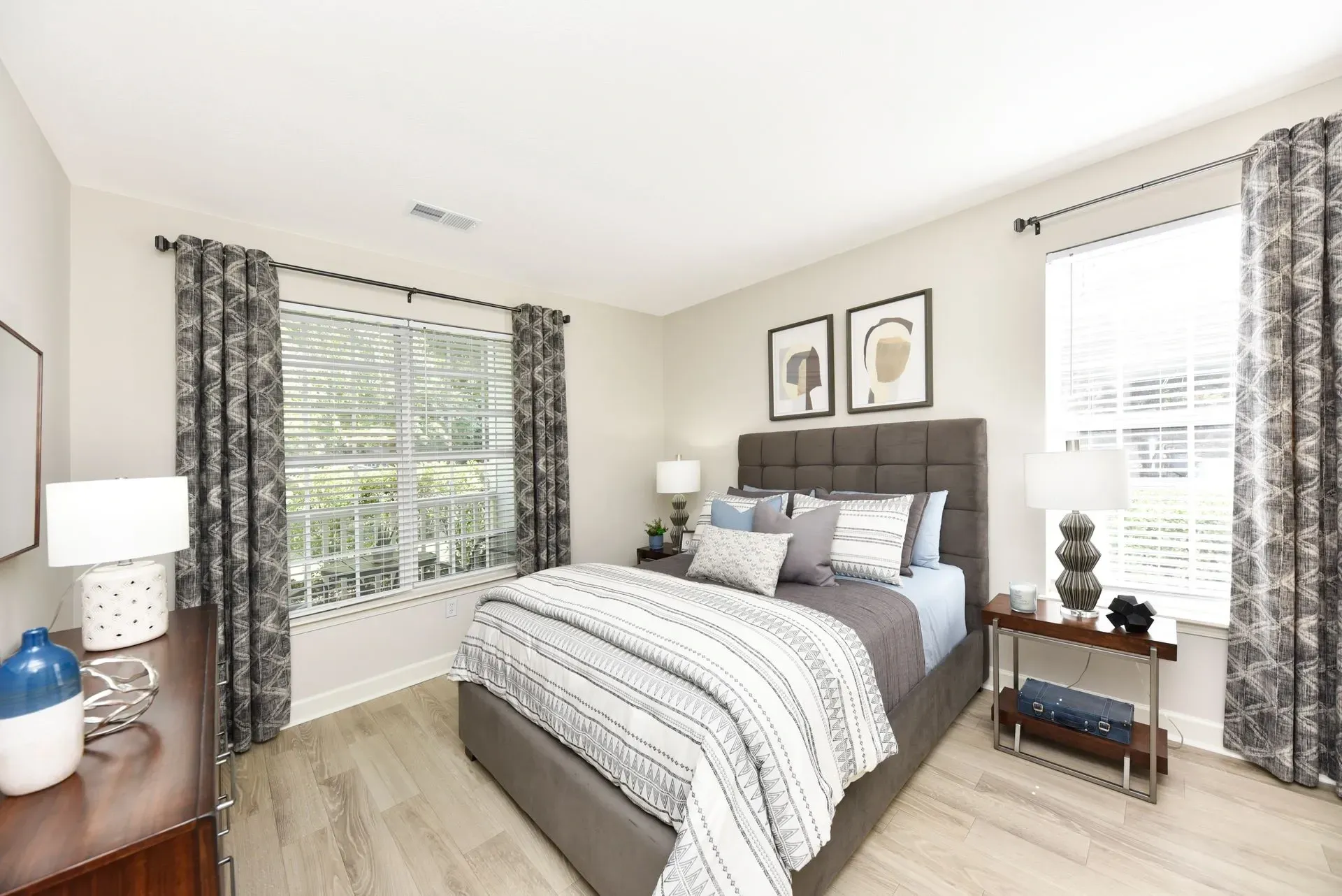 Modern bedroom with a king-sized bed, plush headboard, and decorative pillows. Large windows with patterned curtains offer natural light.