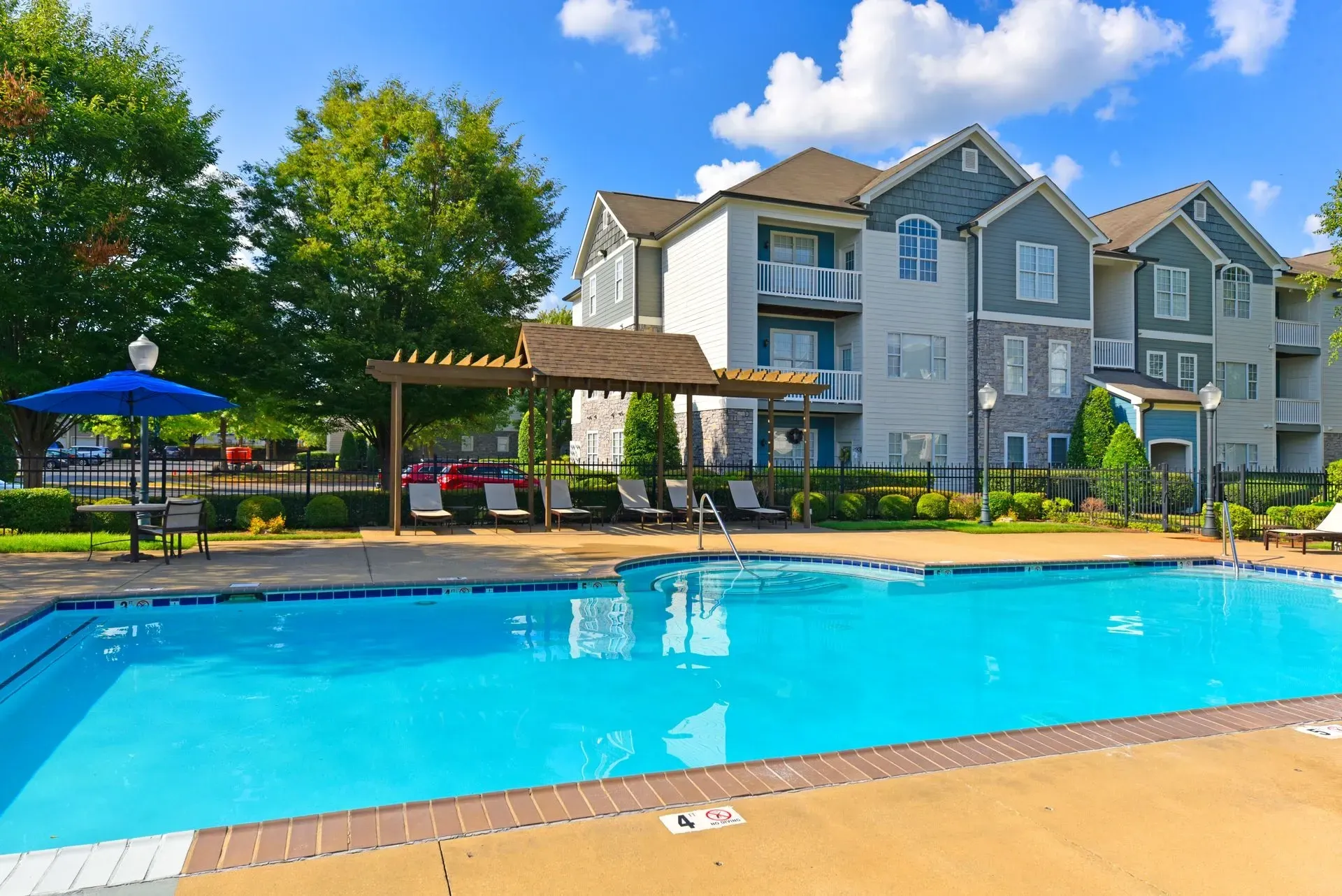 A sparkling blue outdoor swimming pool with lounge chairs and a pergola next to apartment buildings.