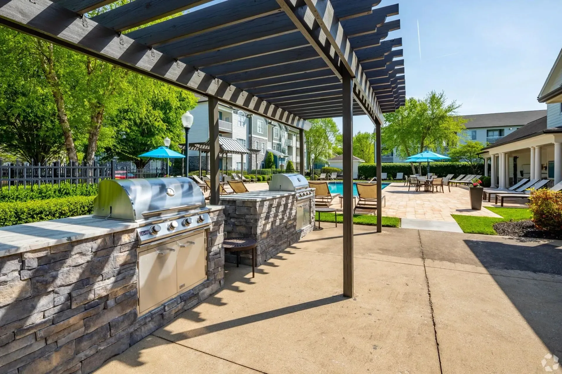 Outdoor grilling station with stainless steel grills and stone base, next to lounge chairs and a swimming pool.