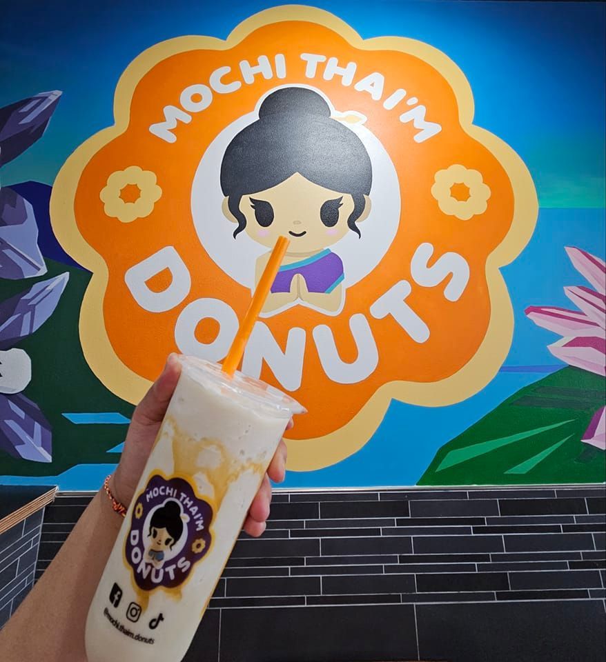Iced drink held in front of a Mochi Thailand Donuts shop sign with a cartoon logo.