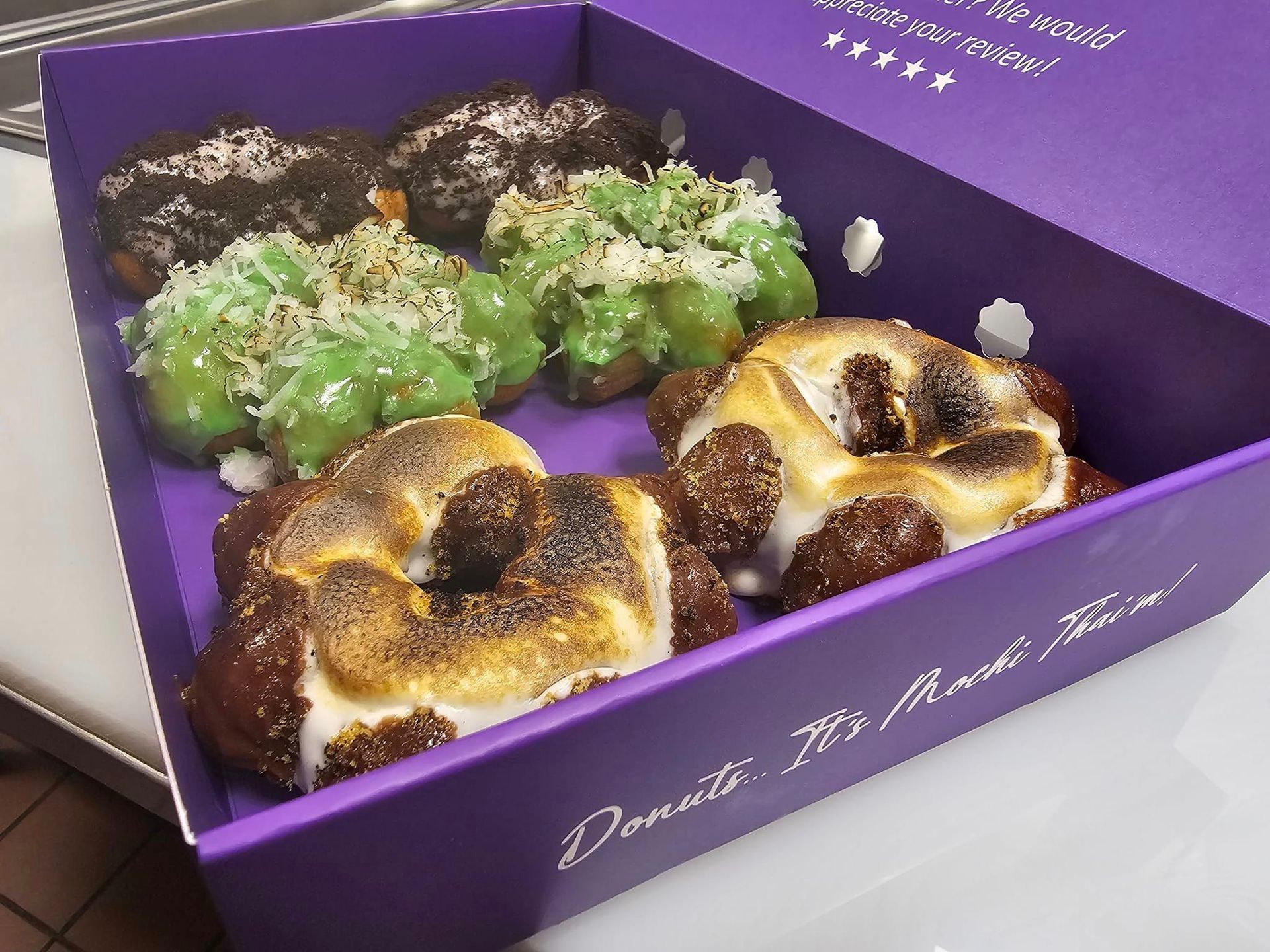 Purple pastry box with assorted glazed donuts, including green and chocolate-topped varieties