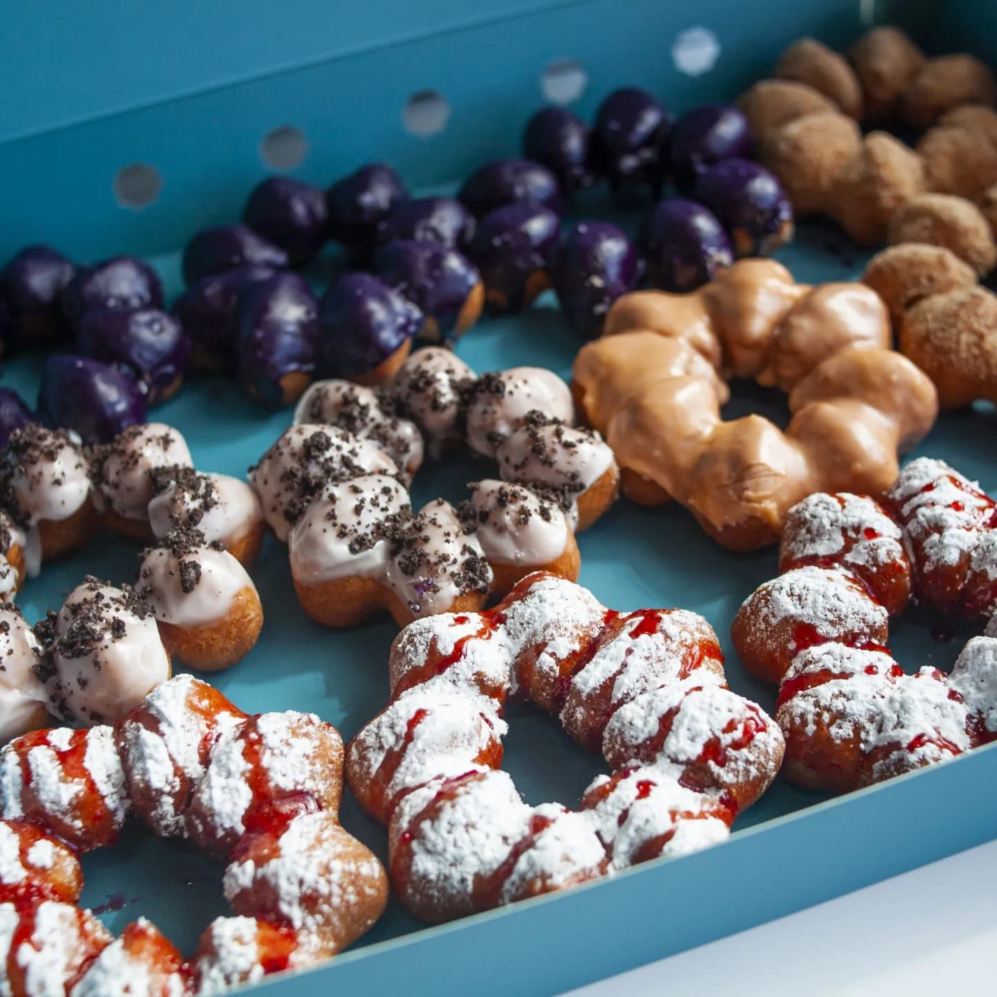 Assorted frosted donuts and pastry bites in a blue box, some drizzled with red icing