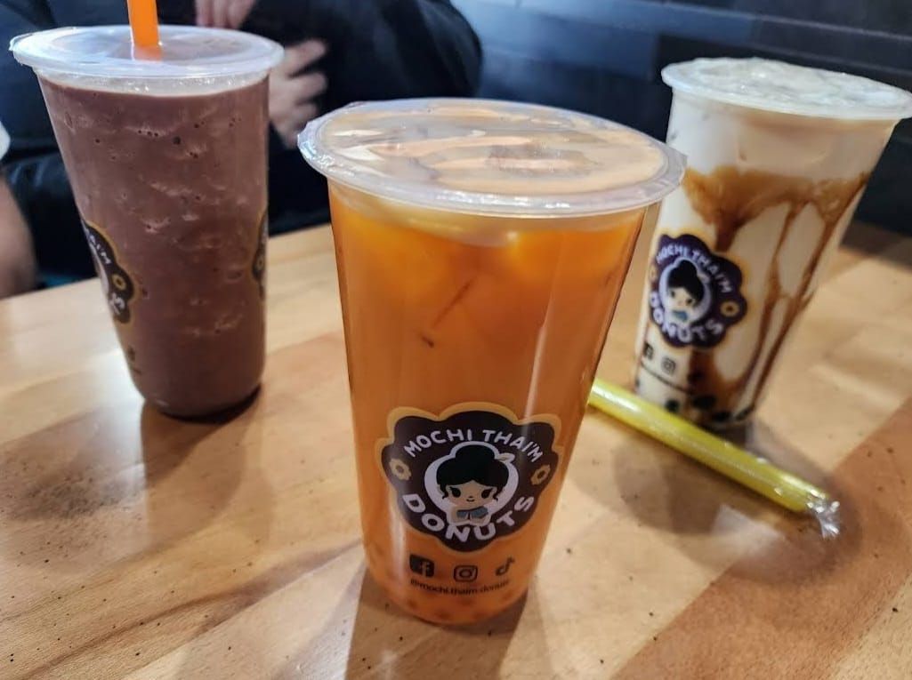 Three iced drinks on a wooden table, including an orange drink in the center and a white drink with chocolate drizzle.