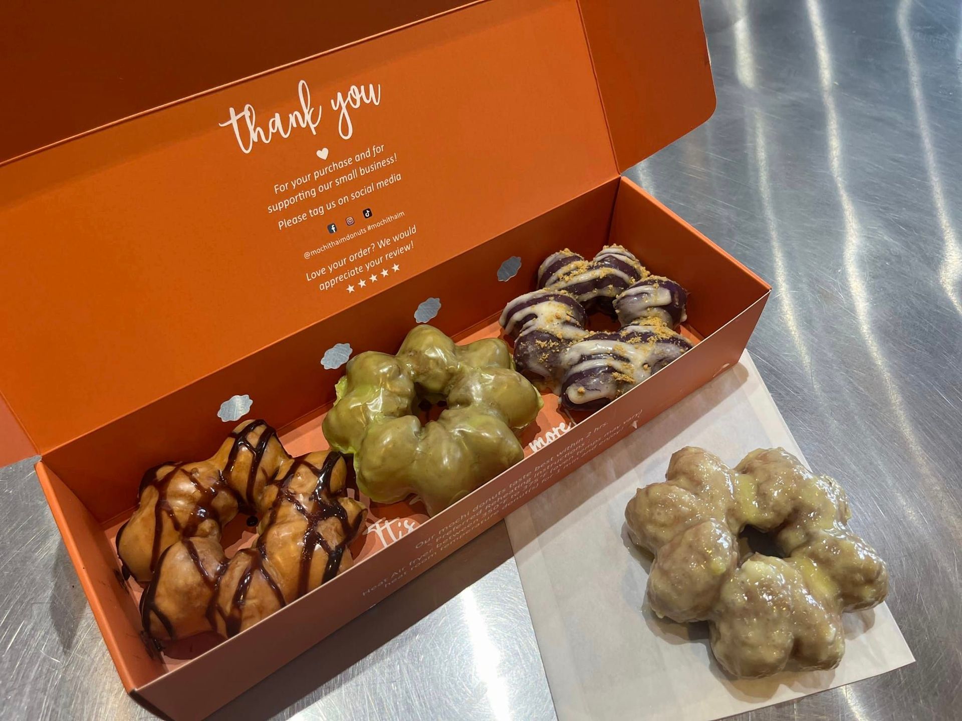 Orange donut box with assorted donuts on a metal surface, plus a small pile of donut holes beside it