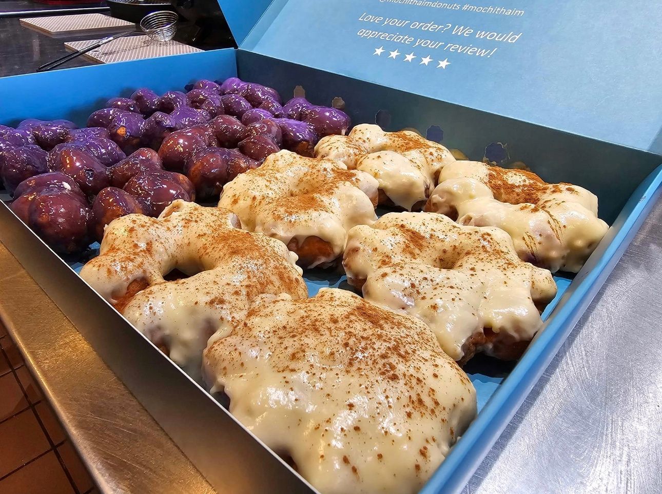Tray of glazed donuts with purple and white icing, sprinkled with cinnamon-like topping in a bakery display box