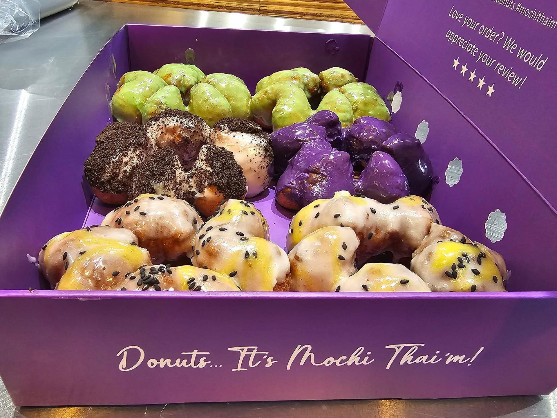 Assorted colorful mochi donuts in a purple box with “Donut. It’s Mochi Thai me!” branding