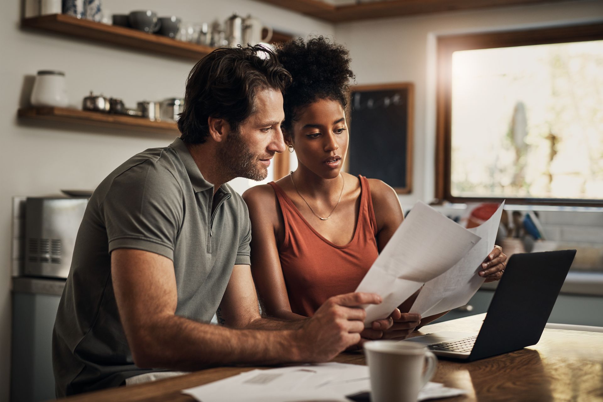 Two people reviewing papers at a kitchen table with a laptop and coffee mug