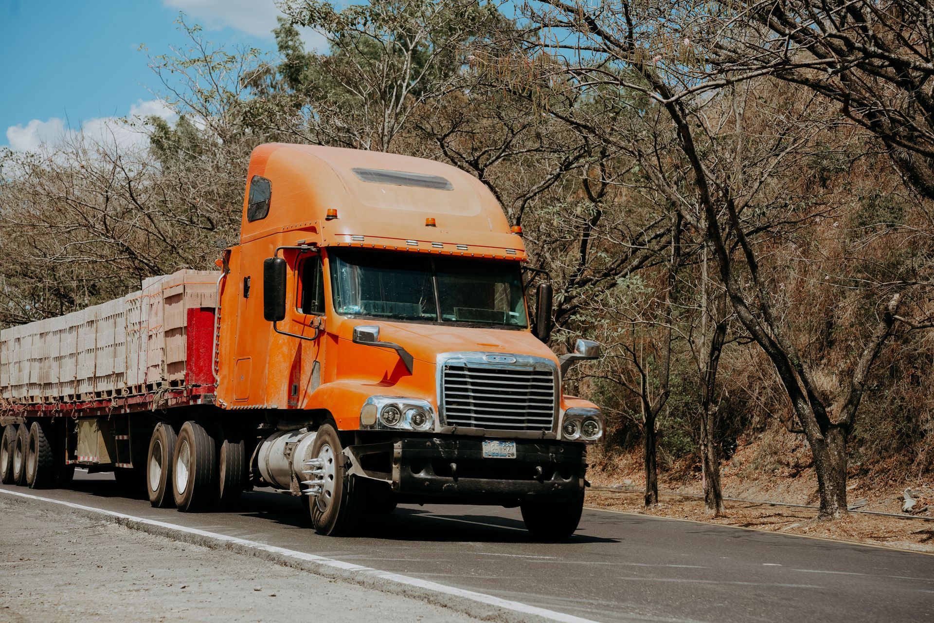 Orange semi-truck hauling a trailer driving on a rural road beside dry trees under a blue sky