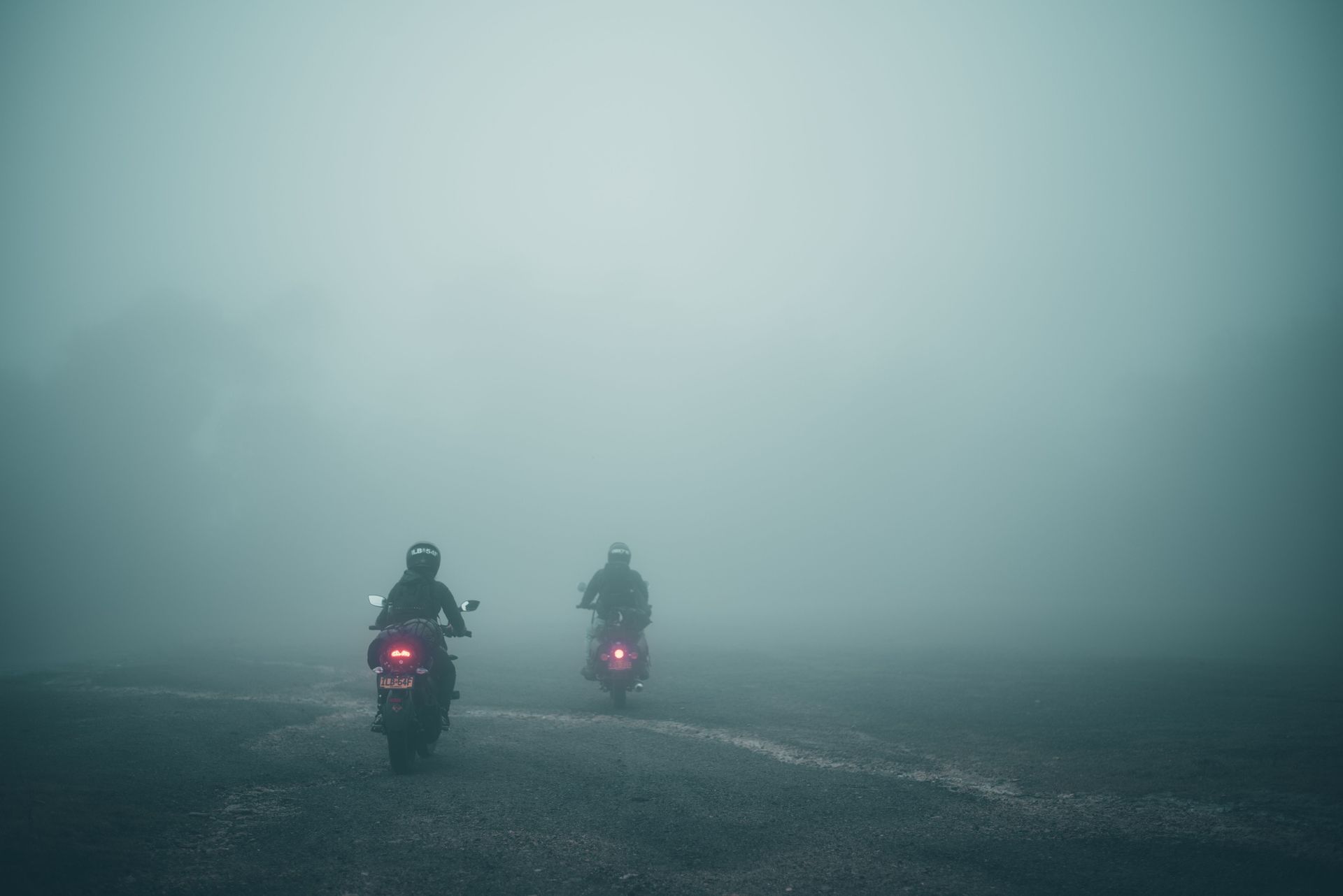 Two motorcyclists ride through dense fog on a wet road, red tail lights glowing.