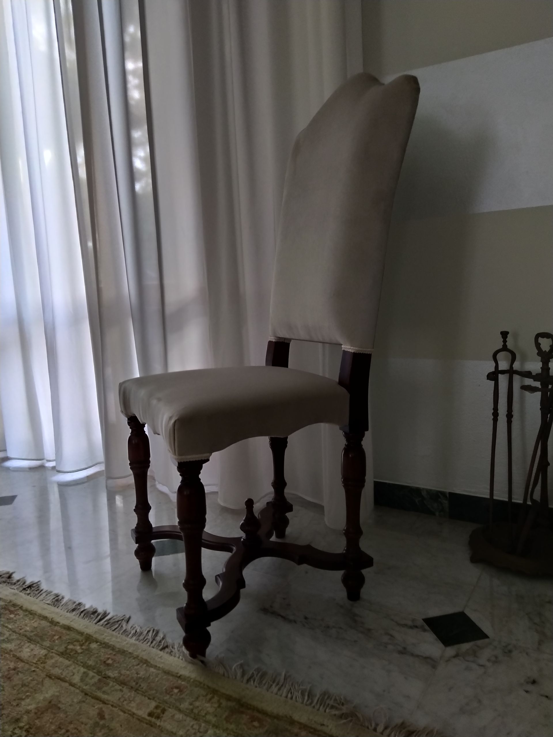 Una sedia da pranzo alta, imbottita di beige, con gambe decorate in legno scuro, posizionata su un pavimento di marmo vicino a tende bianche.