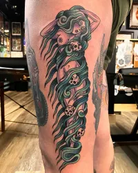 A tattoo on a person's thigh featuring a figure with long, teal, wavy hair intertwined with four small skulls.