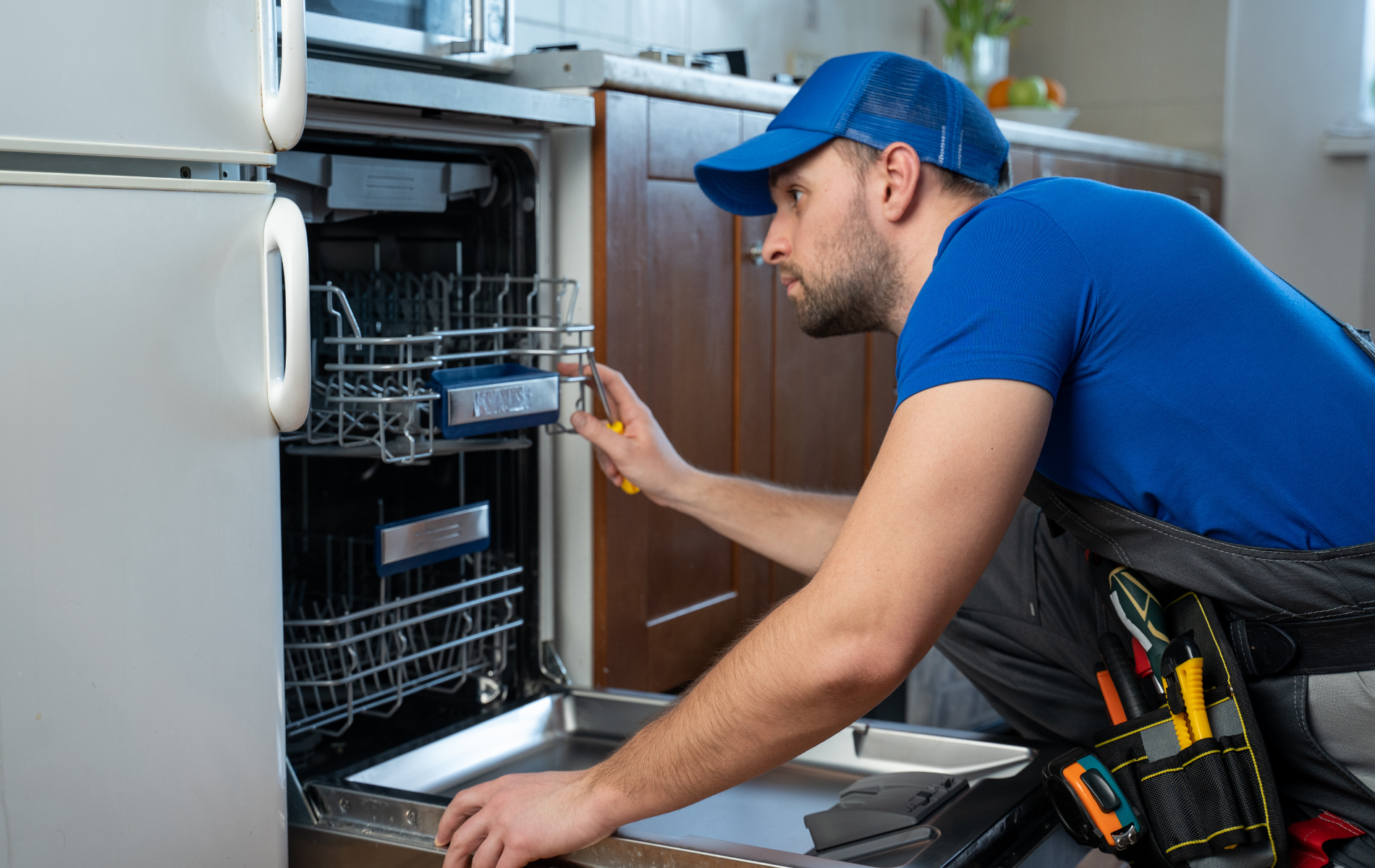 A repairman in blue uniform fixing a dishwasher in a kitchen.