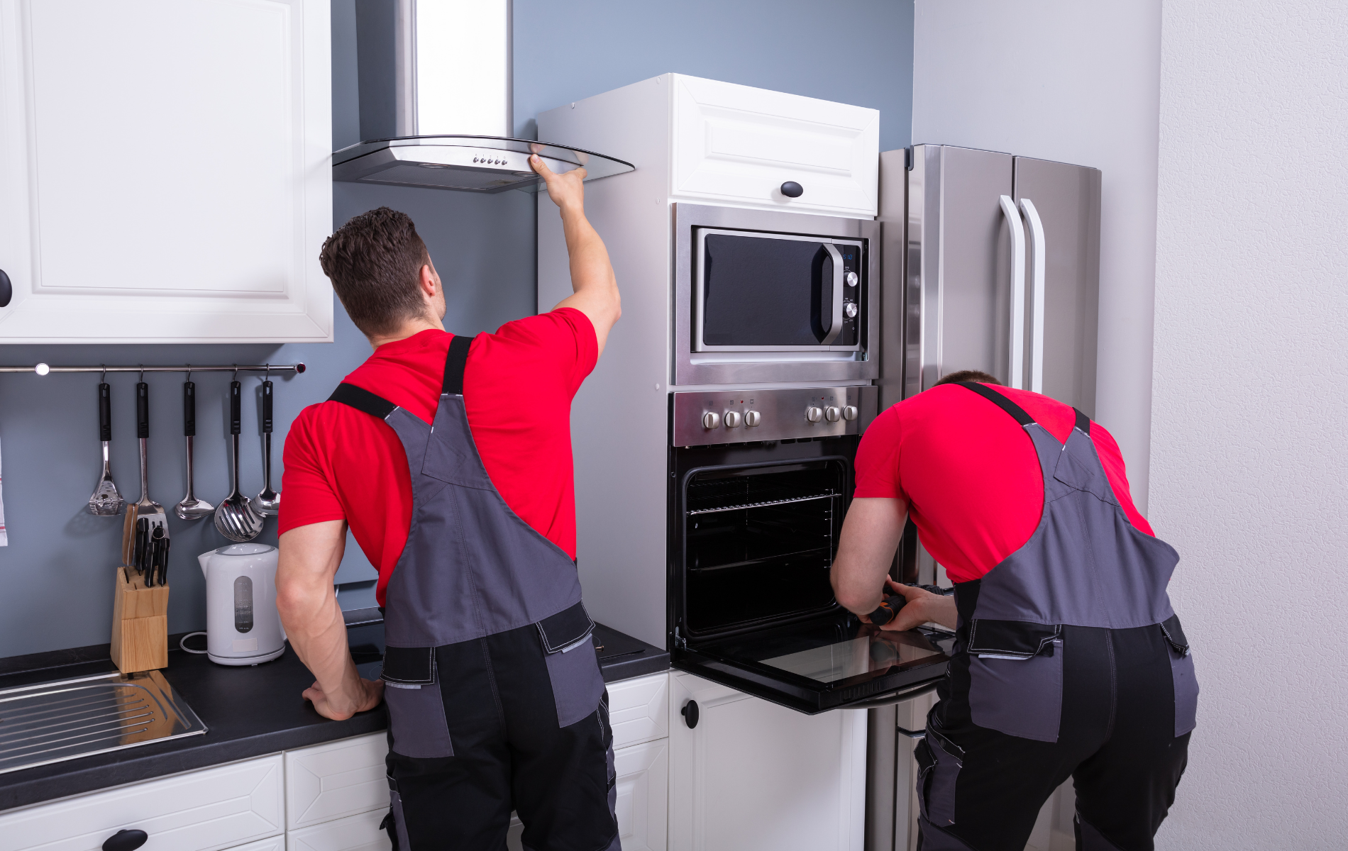 Two appliance repairmen in red shirts and overalls working on a kitchen oven and range hood.