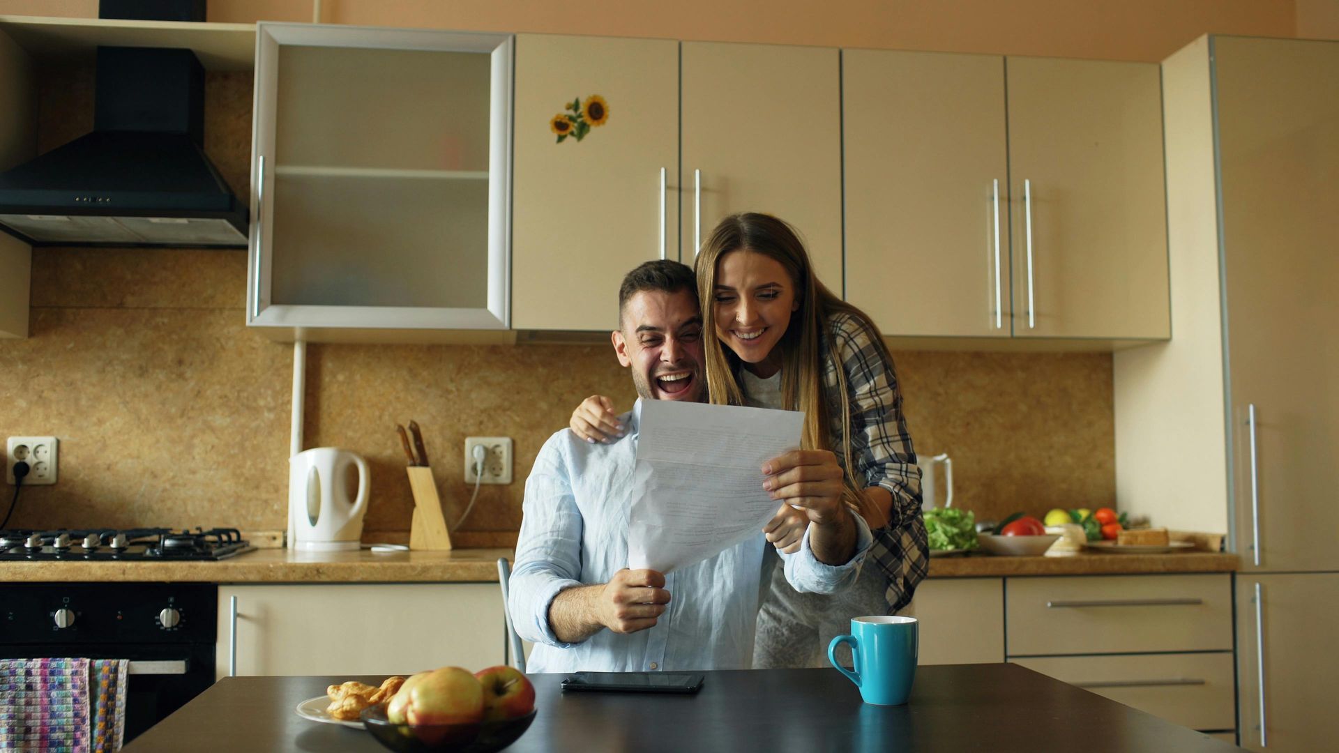 Two people reading a recipe together in a kitchen, with fruit and a mug on the counter.