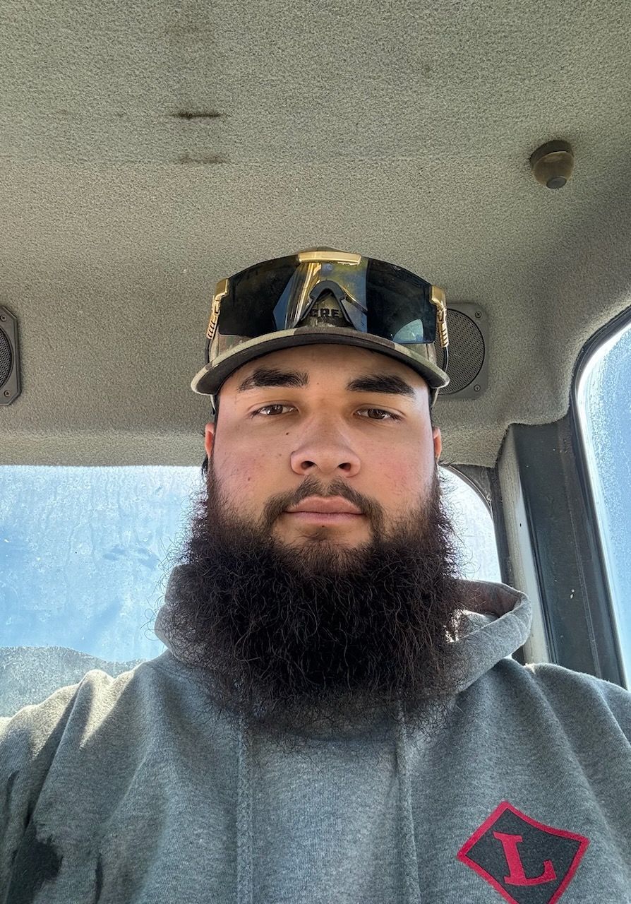 Moko with a beard wearing a hat and sunglasses is sitting in a car.