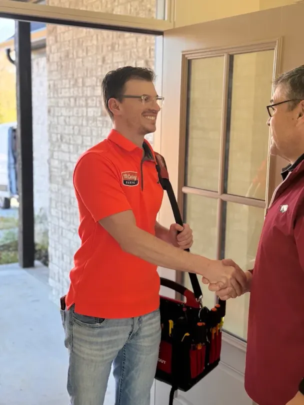Two men shake hands in a doorway, one in a red shirt with a tool bag and one in a orange shirt.