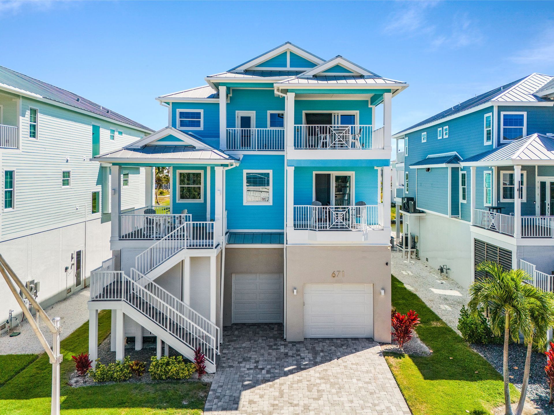 Bright blue elevated beach house with white trim, front stairs, and a garage, between neighboring homes.