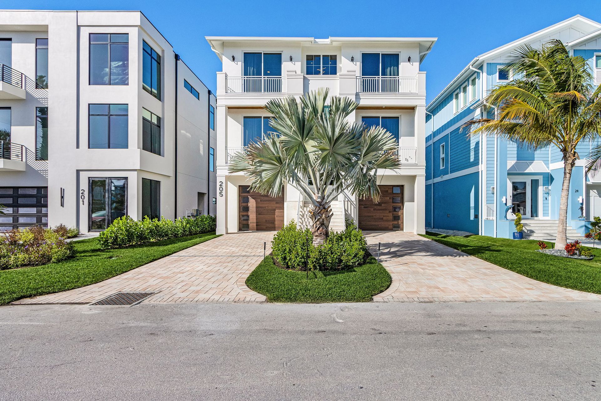 Three-story white house with balconies, two car garage, and palm tree in front, next to other houses.