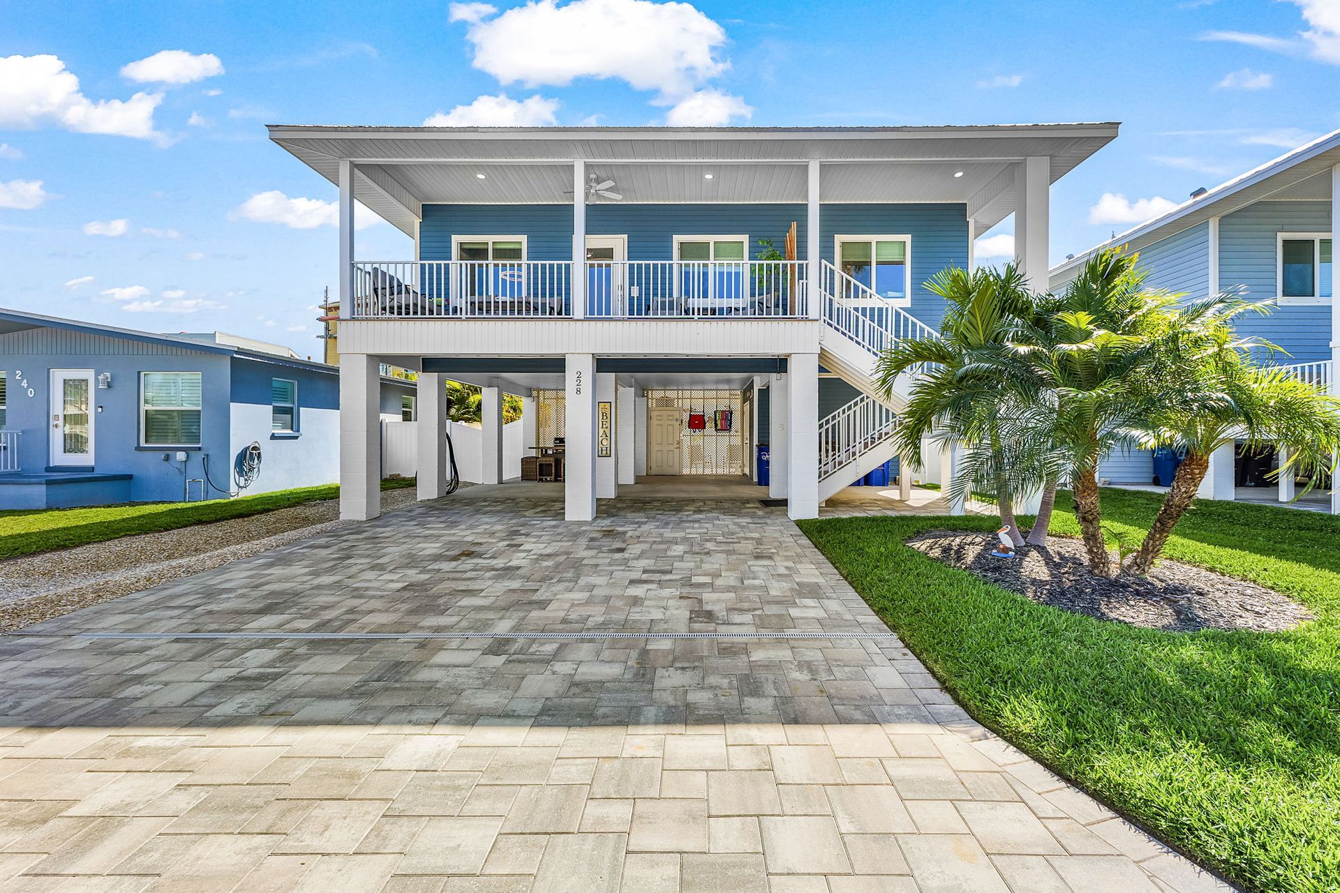 Modern two-story house with a stone driveway, balcony, and palm tree under a blue sky