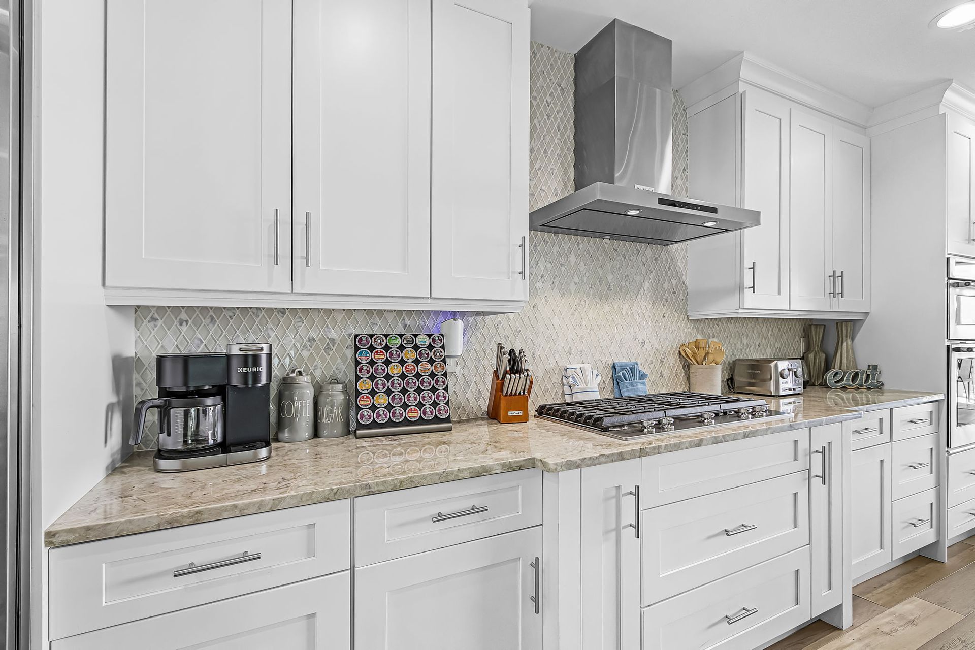 Bright white kitchen with marble counters, white cabinets, coffee maker, stove hood, and decorative backsplash