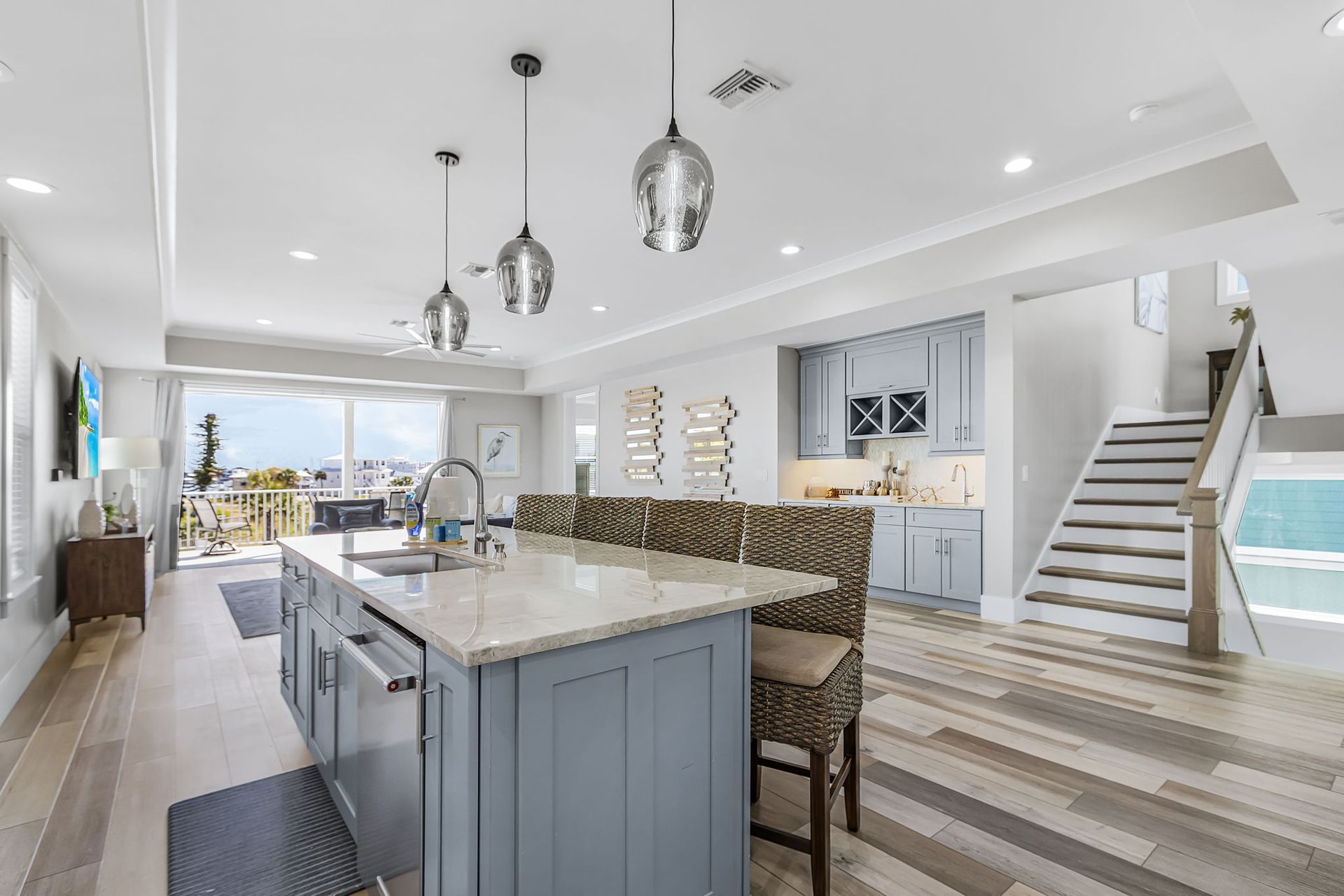 Bright modern kitchen and living space with island, pendant lights, staircase, and ocean view.