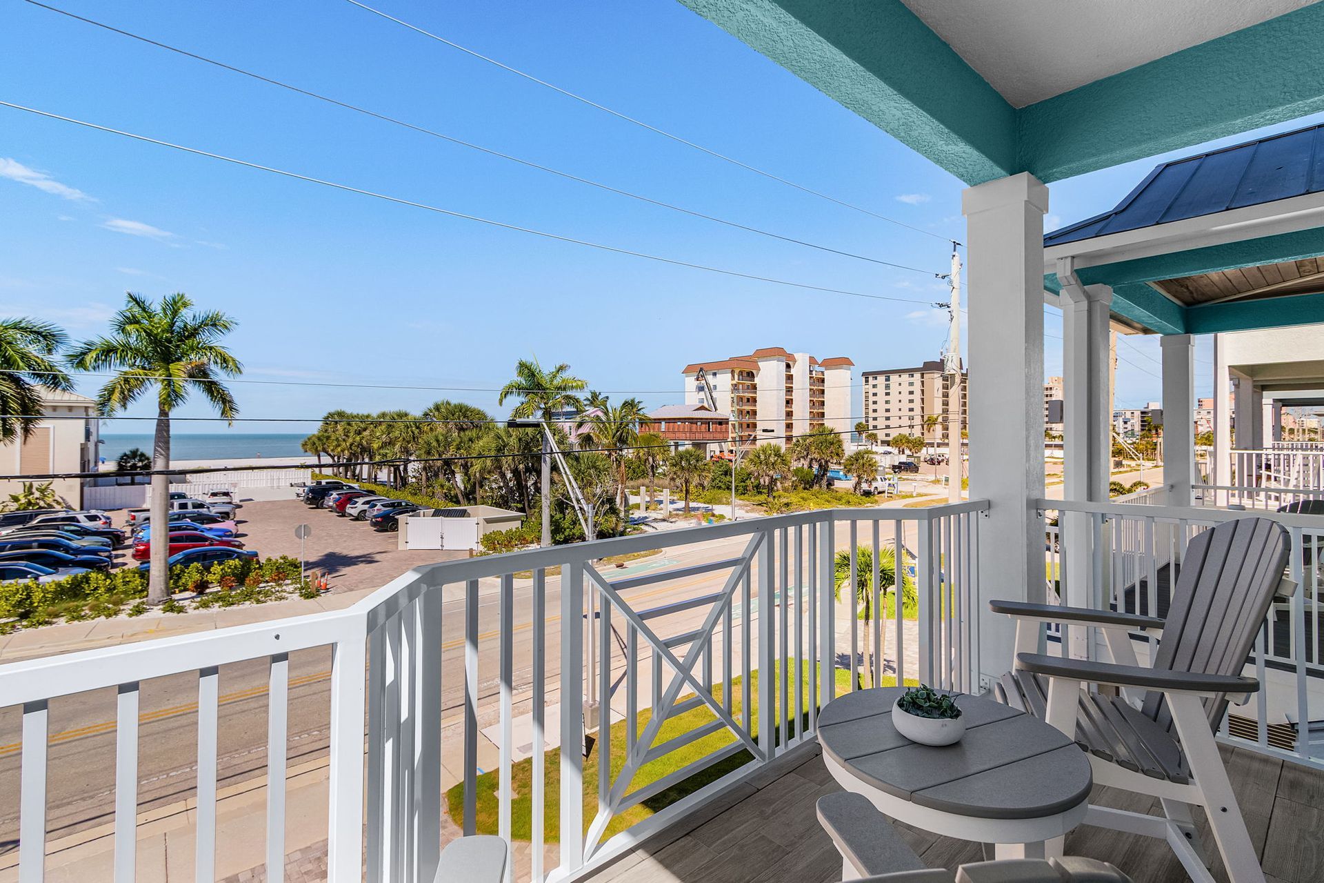 Balcony with white railing and chairs overlooking a sunny beach, palm trees, and seaside buildings