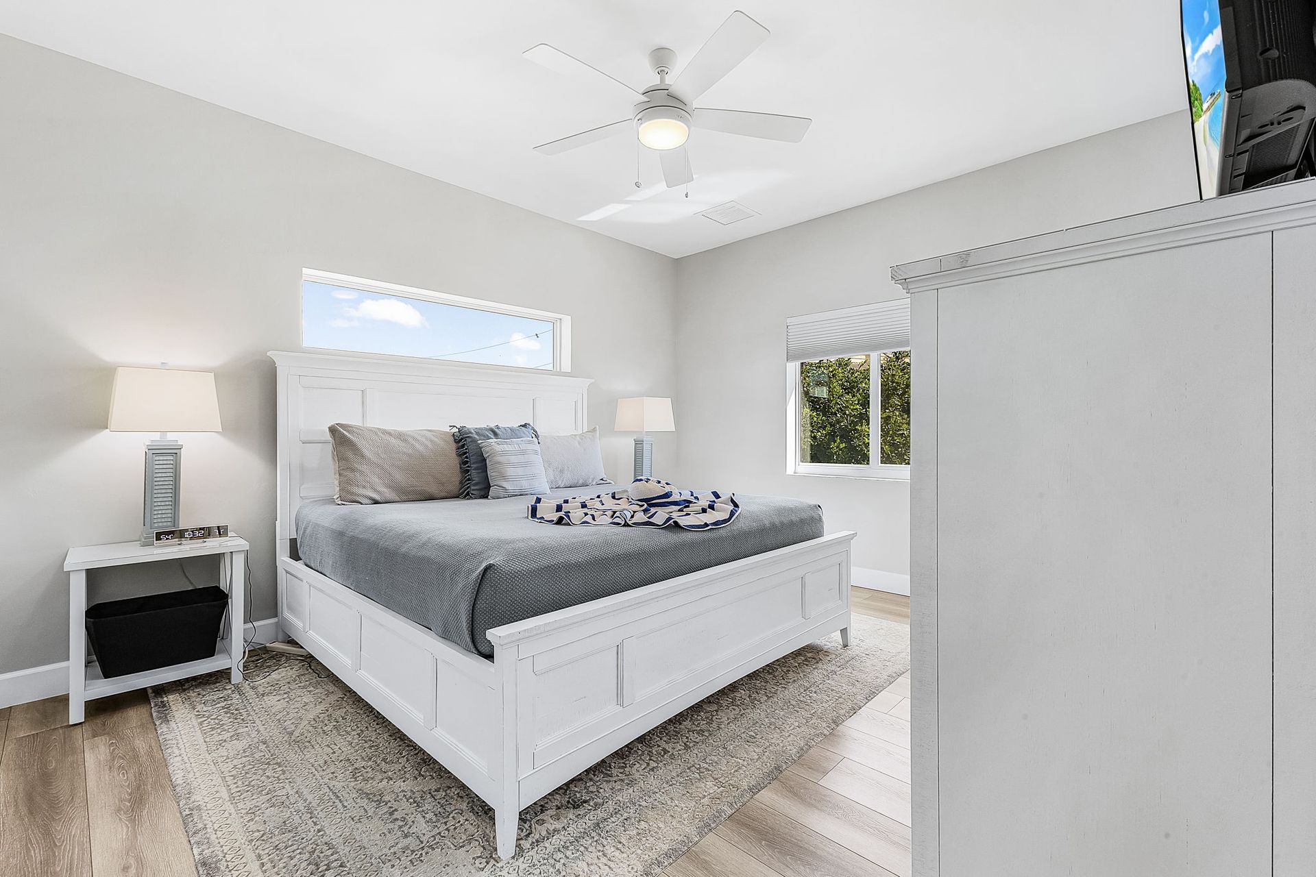 Bright bedroom with a white bed, gray bedding, nightstand, ceiling fan, and large windows.