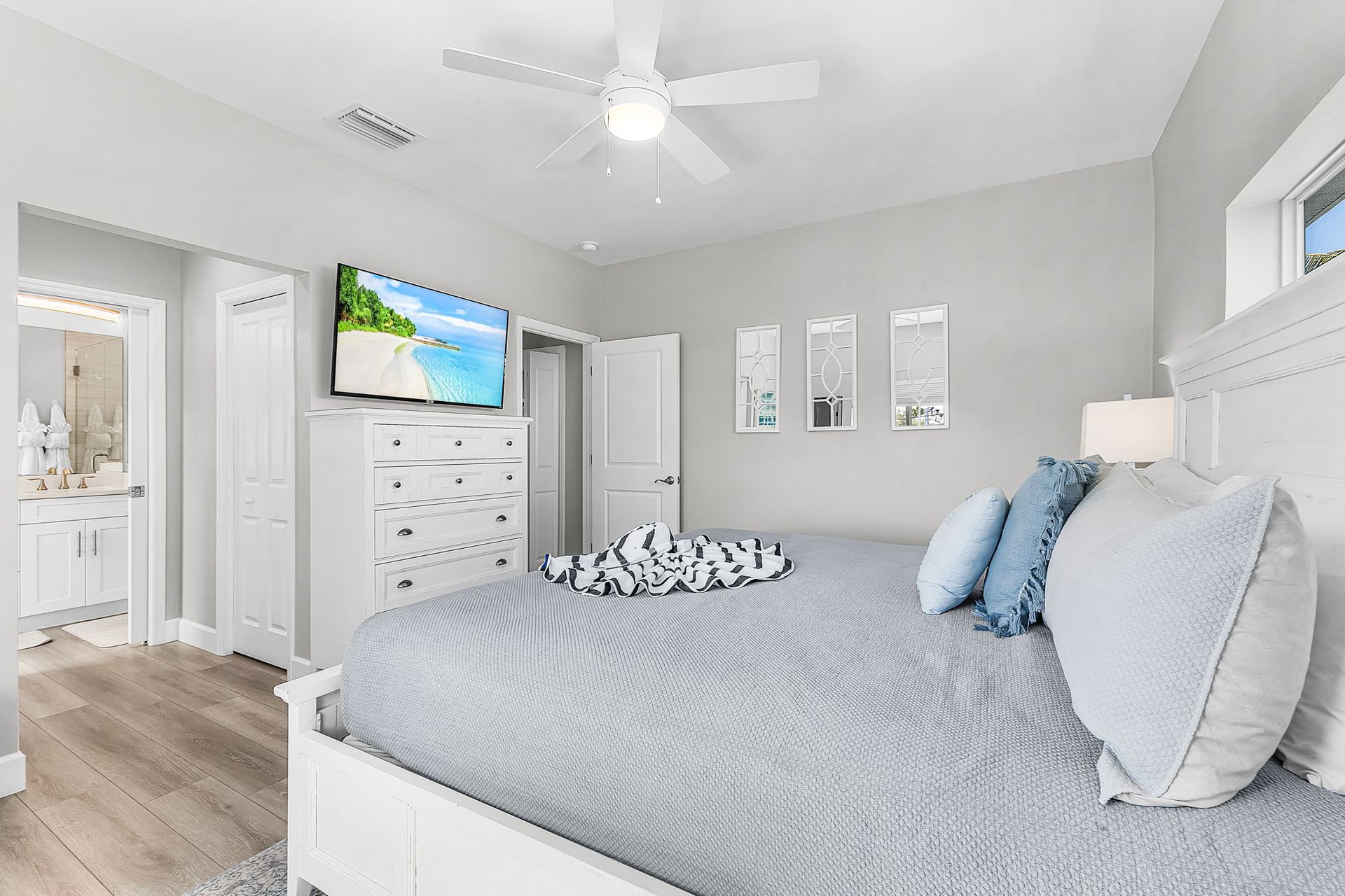 Bright gray bedroom with a bed, dresser, wall-mounted TV, ceiling fan, and beach artwork.