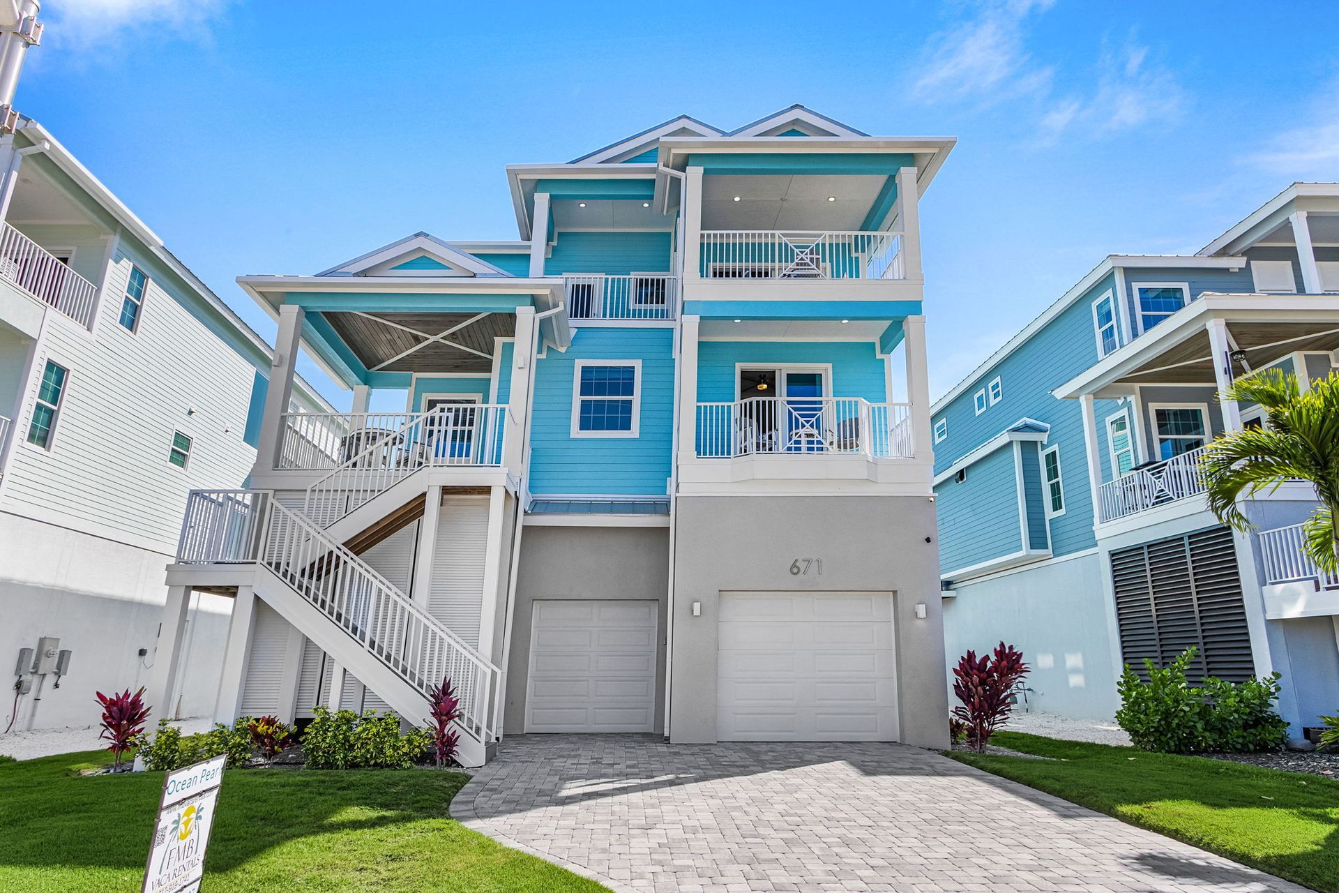 Blue two-story beach house with white trim, balconies, stairs, and garage on a sunny street