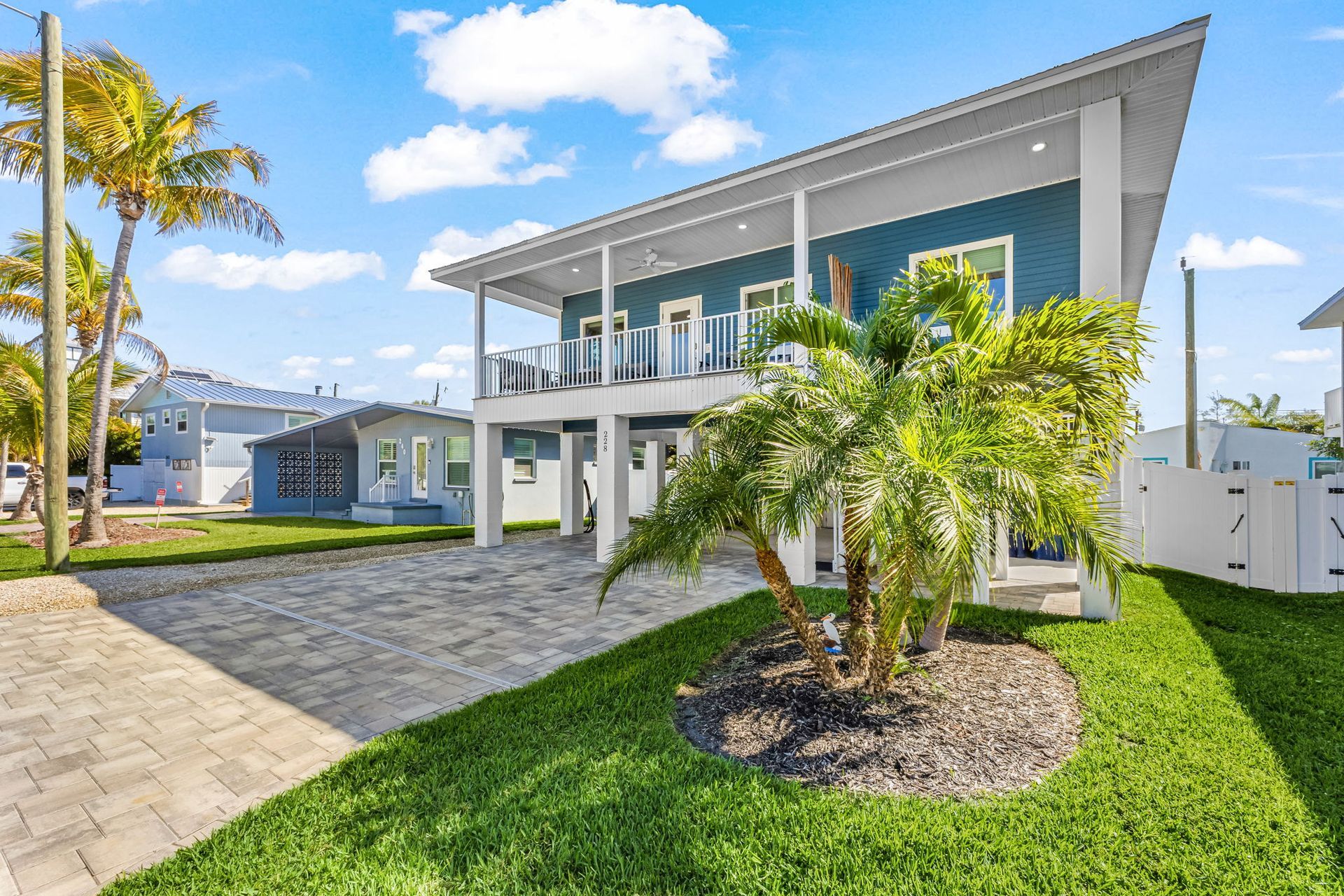 Modern two-story house with white trim, blue siding, palm trees, and a landscaped driveway in bright sunlight