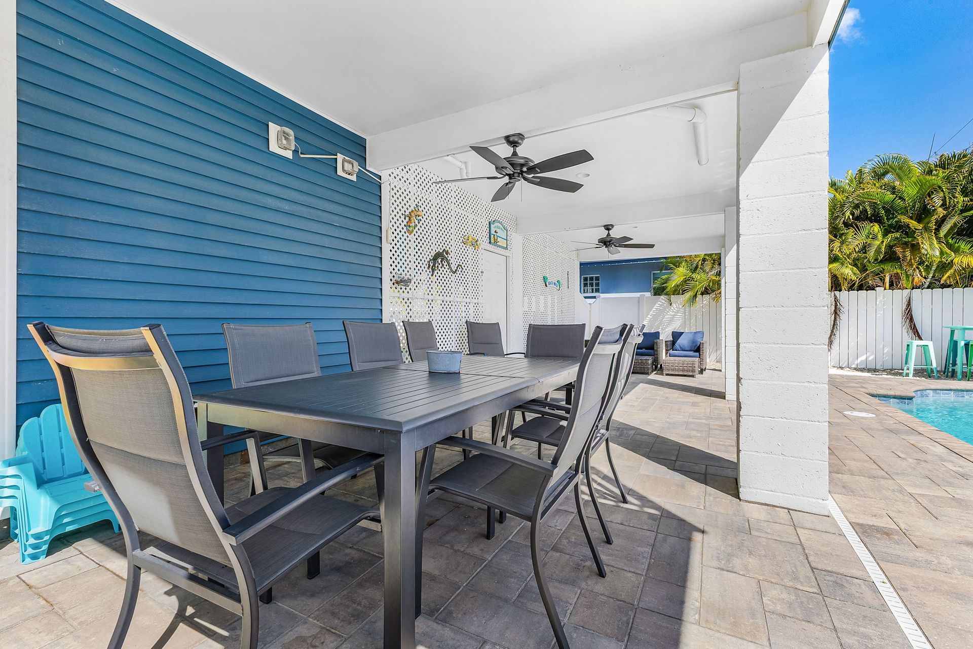 Covered patio with gray dining table and chairs beside a blue wall, leading to a pool area.