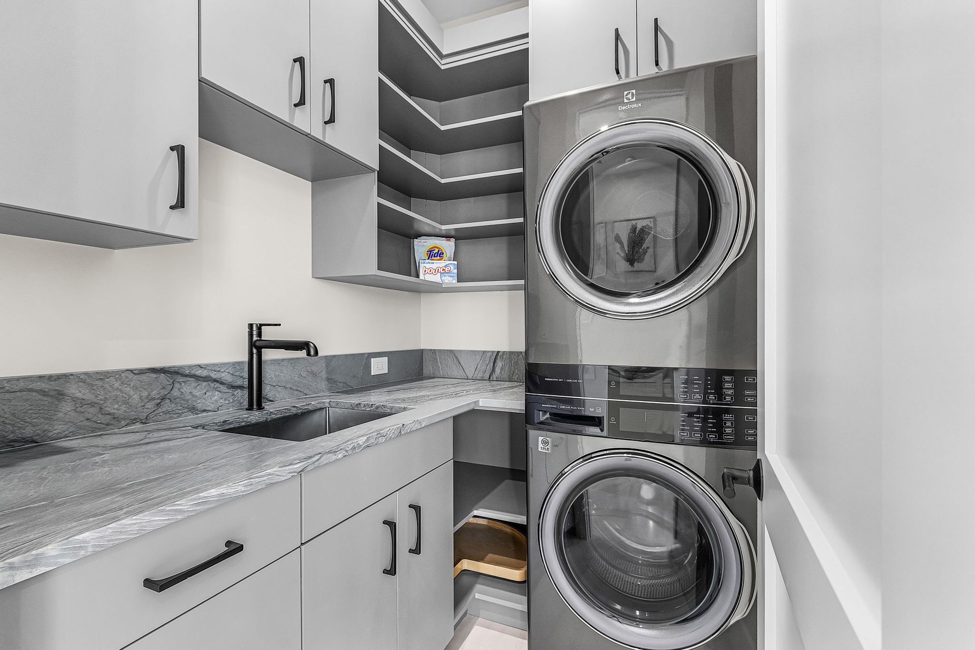 Gray laundry room with stacked washer and dryer, cabinets, counter, and sink.