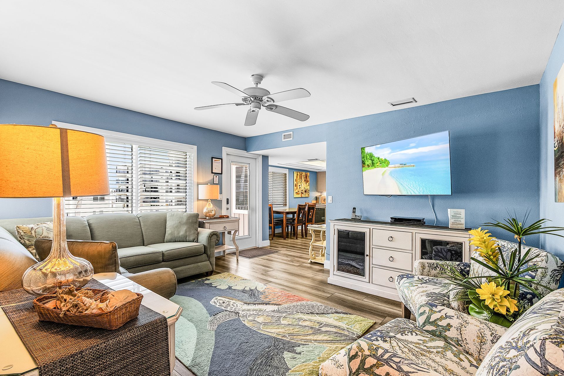 Bright living room with blue walls, white sofa, TV console, lamp, and patterned rug, leading to a dining area.