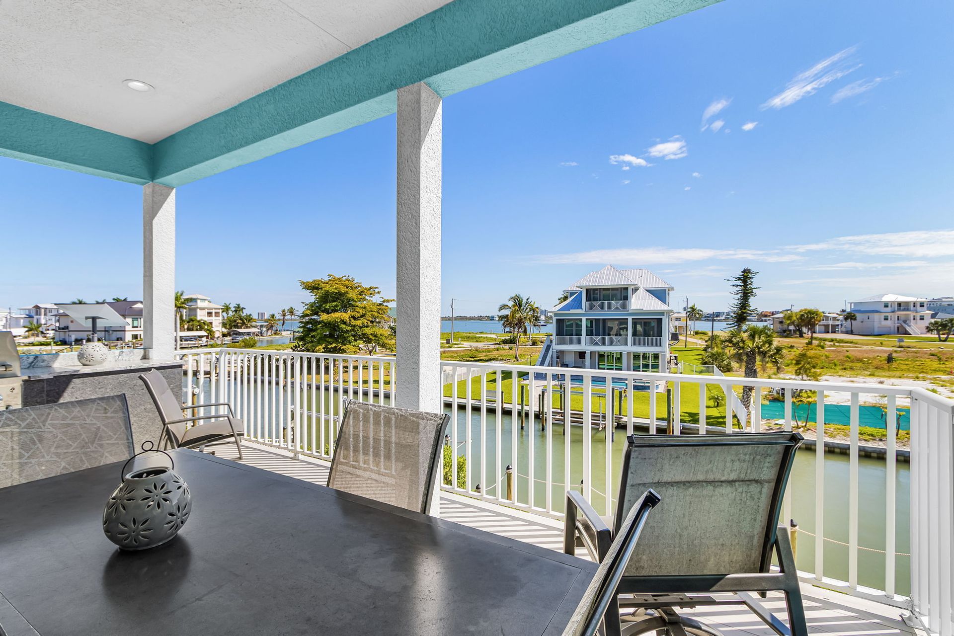 Balcony with chairs and a small table overlooking a sunny waterfront neighborhood and pool