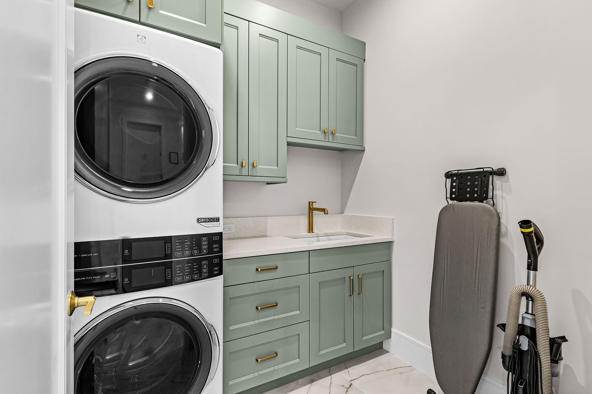 Laundry room with stacked washer and dryer, sage green cabinets, and ironing board.